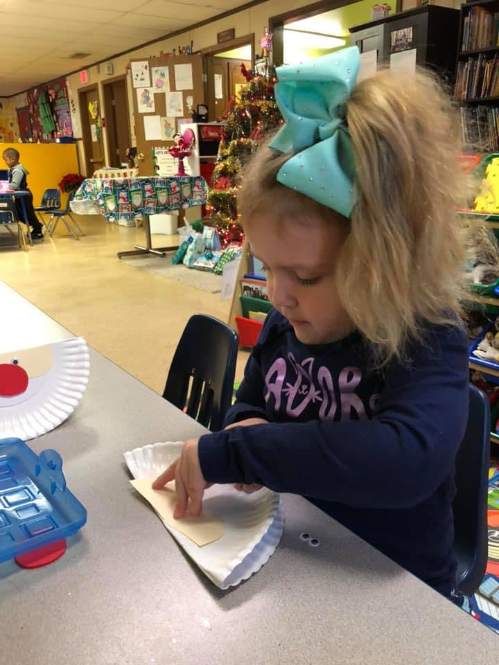 Child with a large bow in their hair working on a craft at a table in a classroom