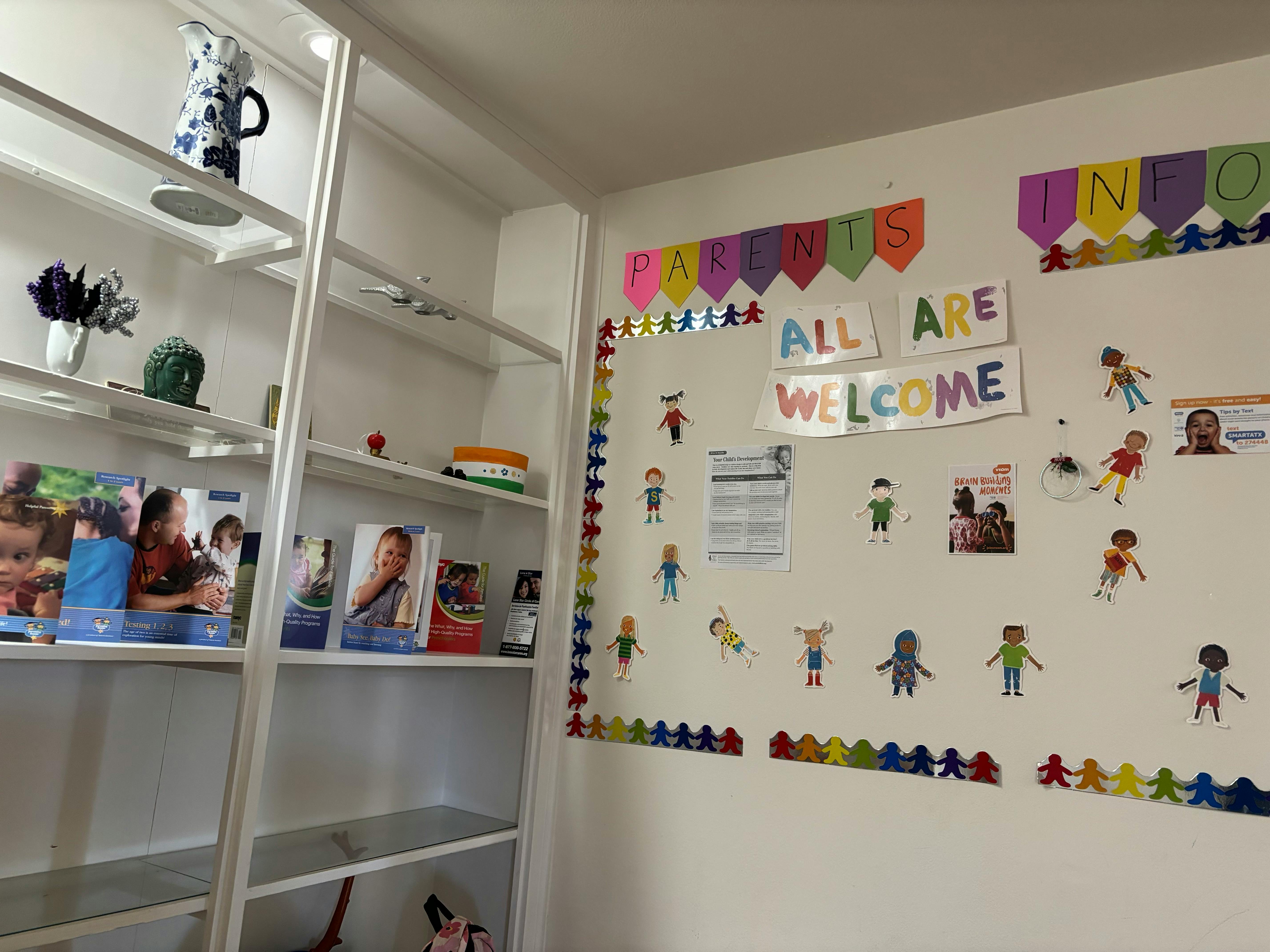Wall with "All Are Welcome" sign and paper cutouts next to a white bookshelf