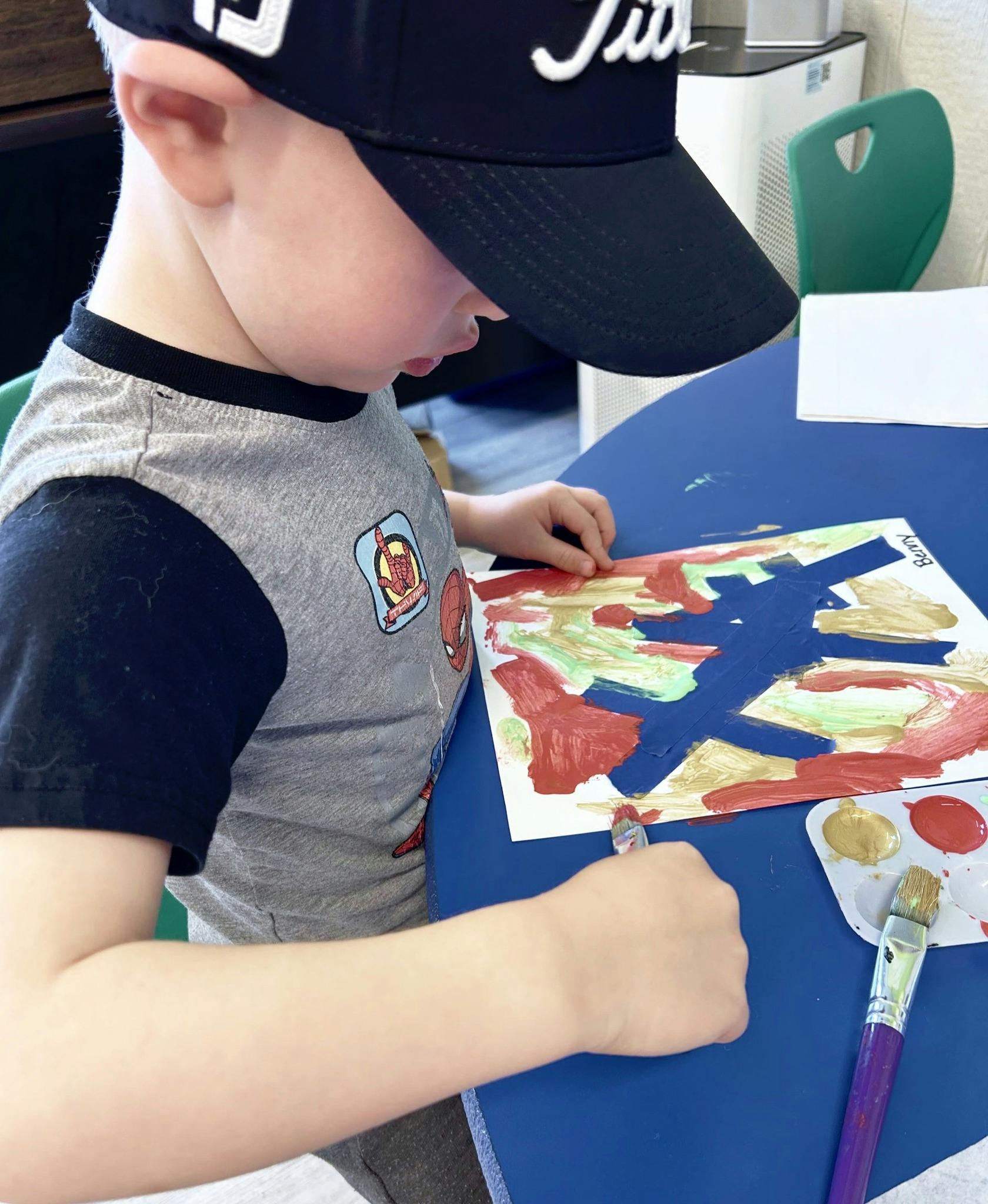 Child in a baseball cap painting on paper at a blue table
