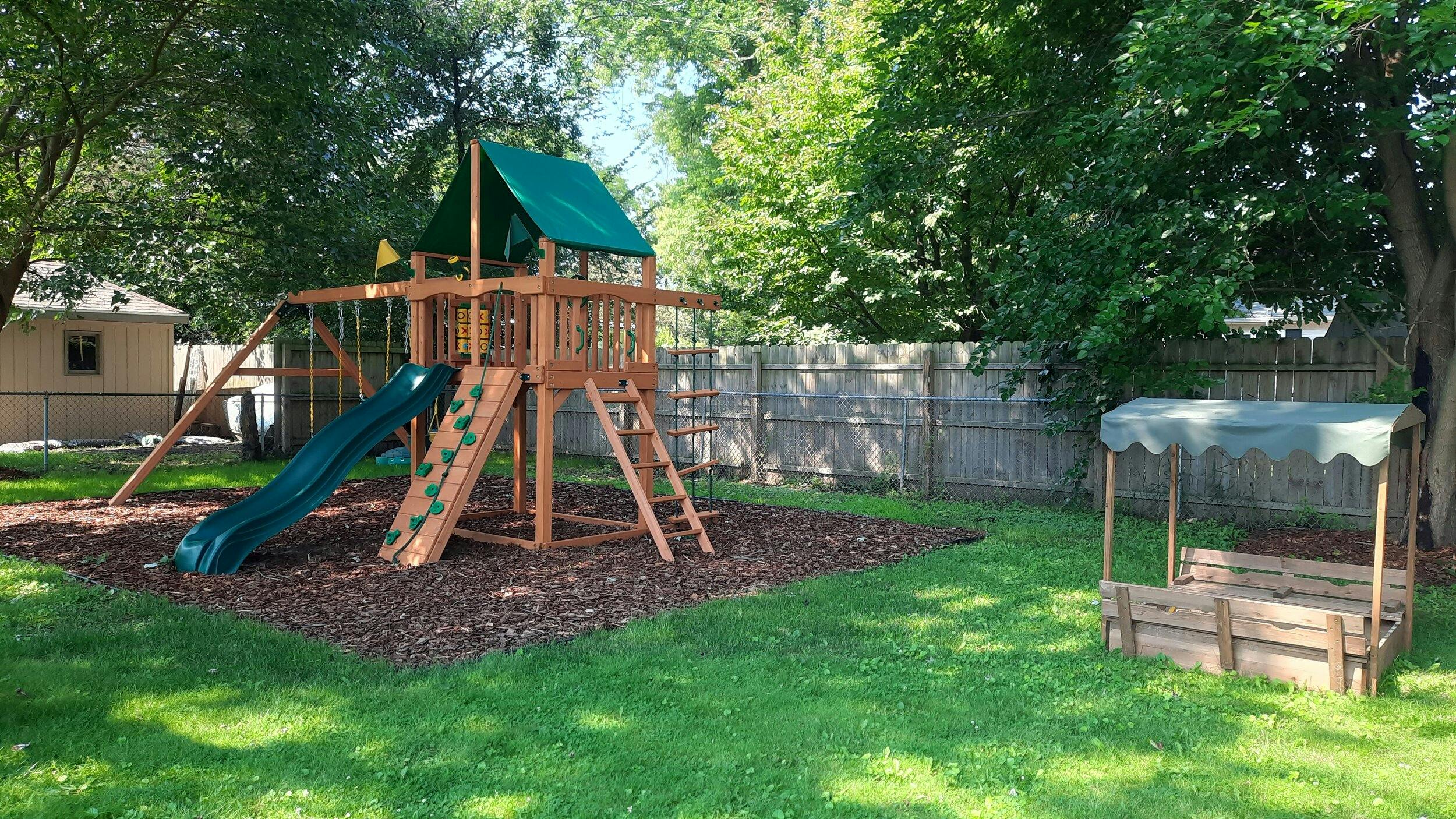 Wooden playset with slide and swings next to a sandbox in a grassy yard