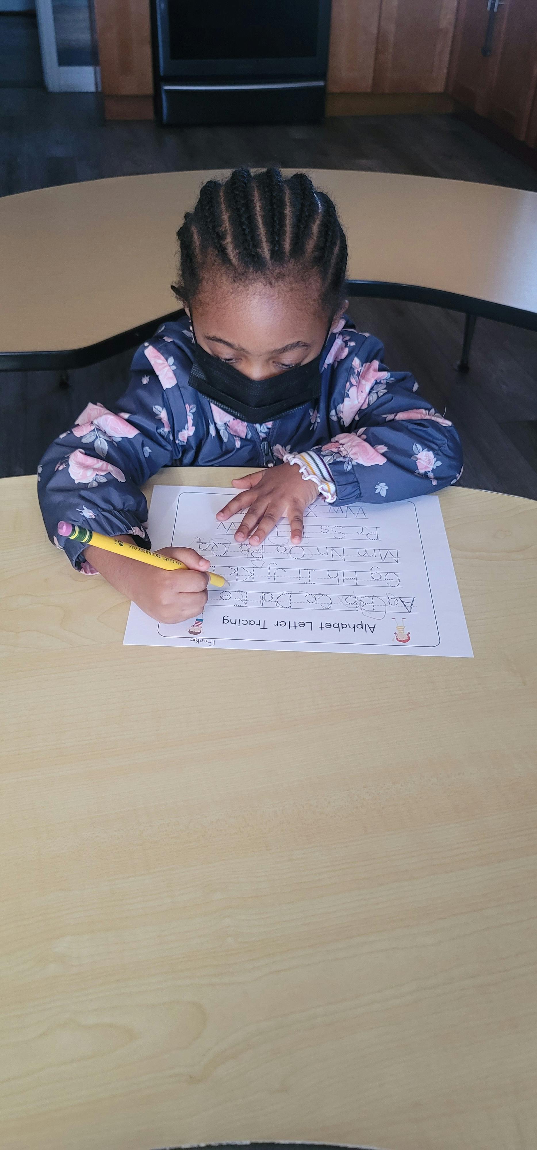 Child in a face mask tracing letters on a worksheet with a pencil