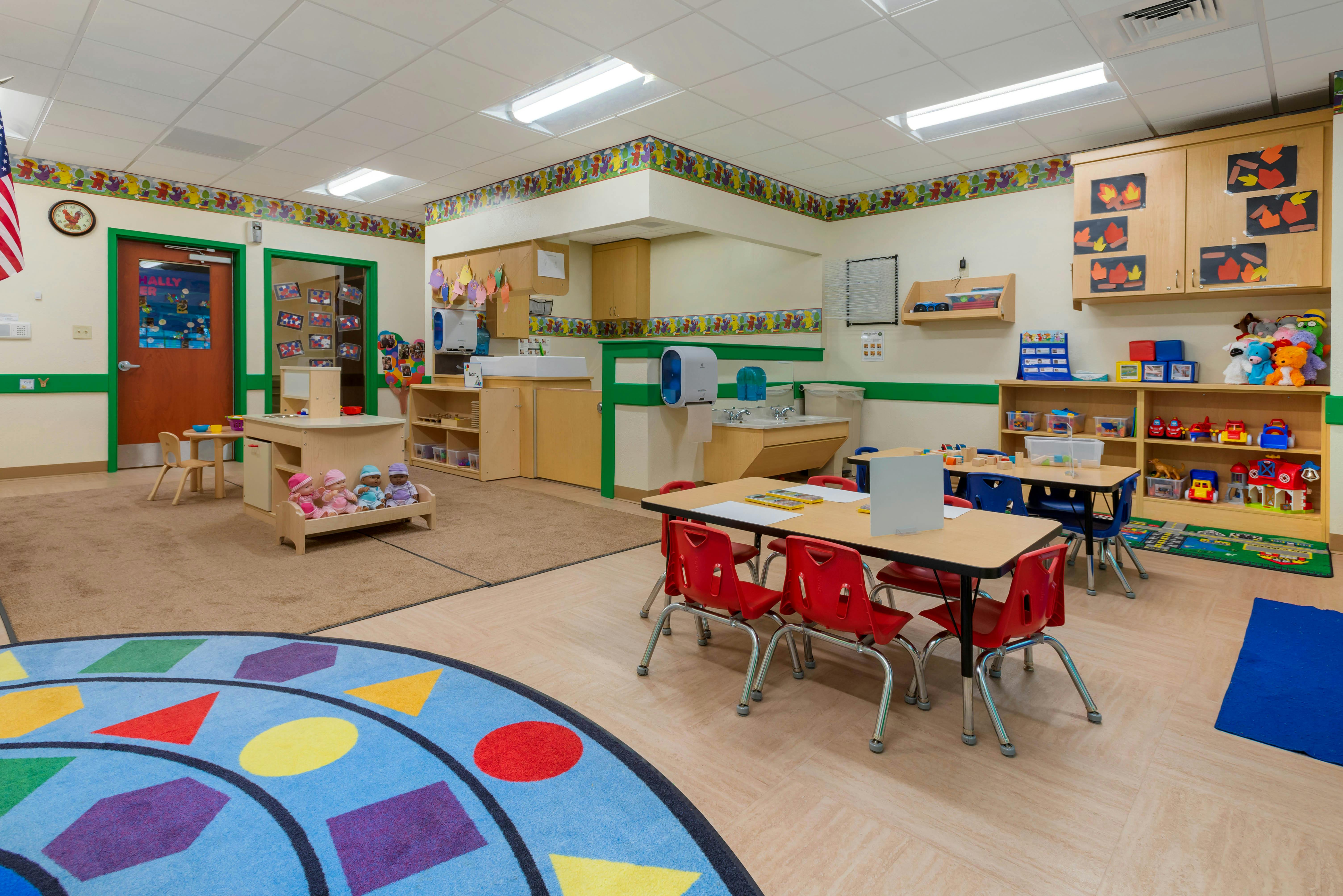 Colorful classroom with child-sized tables and chairs, play area with dolls, and sink