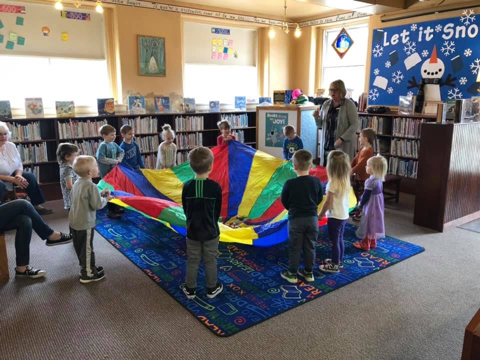 Children and adults playing with a colorful parachute in a room with bookshelves