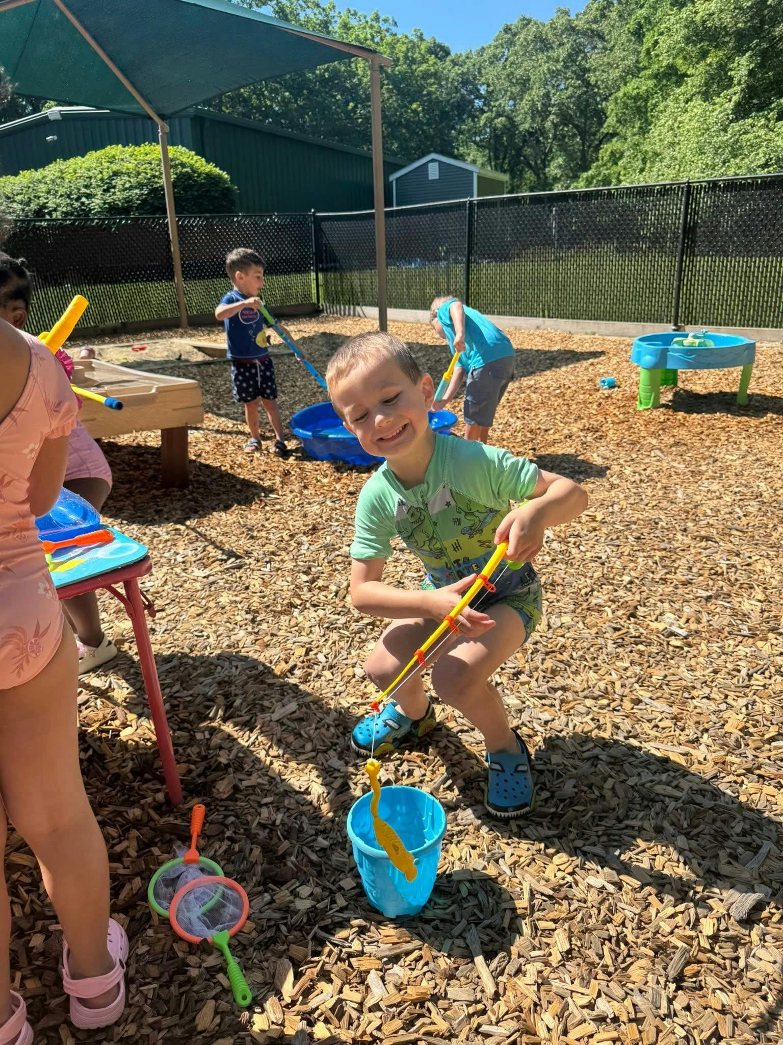 Child playing with a toy fishing rod and blue bucket on a wood chip playground