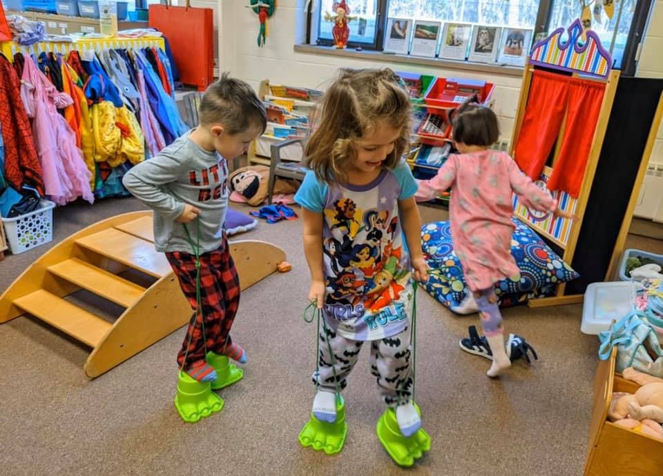 Two children walking on green monster feet stilts in a classroom