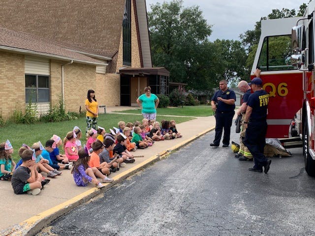 Children sitting on a sidewalk listening to firefighters next to a fire truck