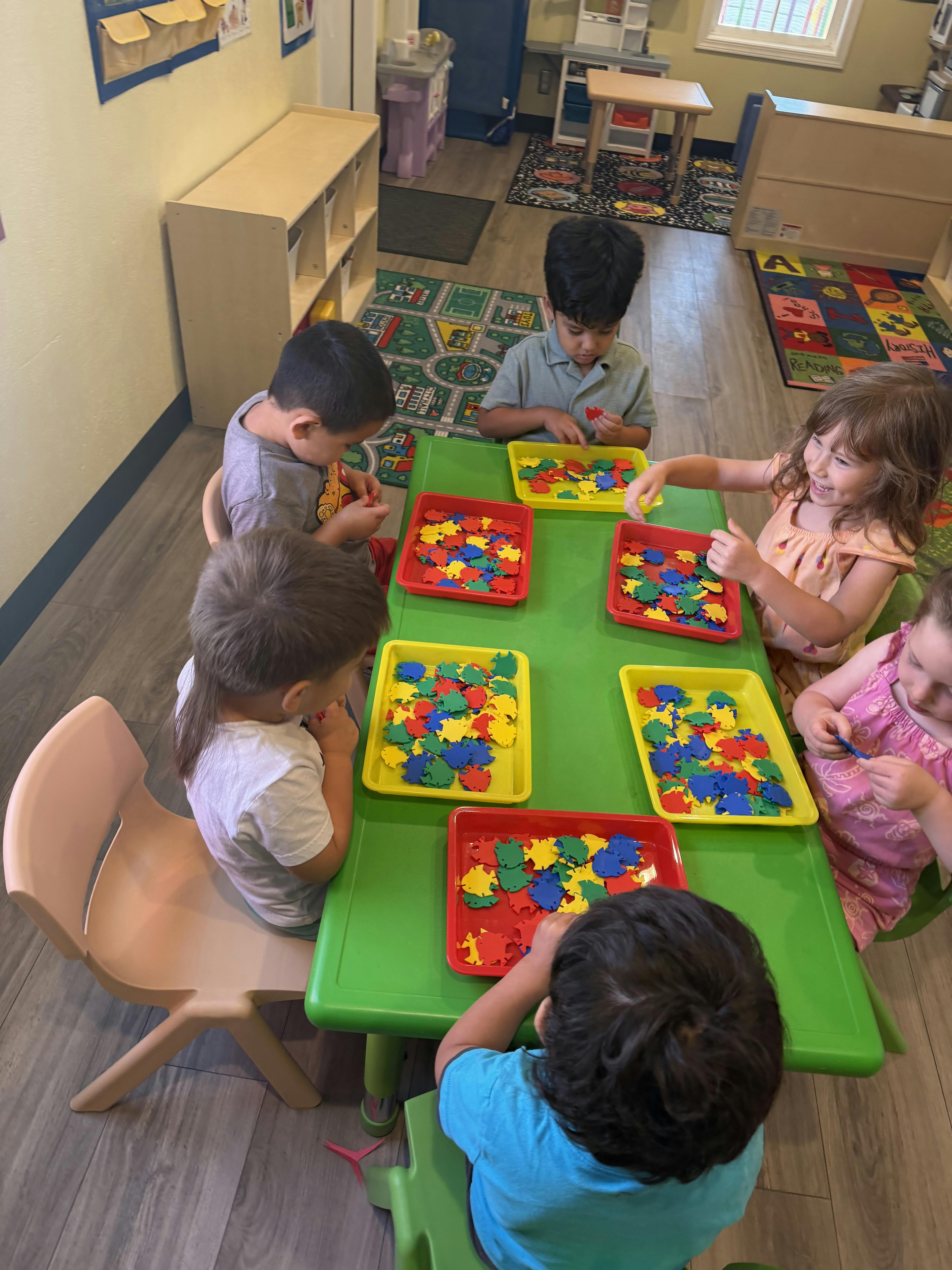 Children sitting at a table in a classroom sorting colorful shapes into trays