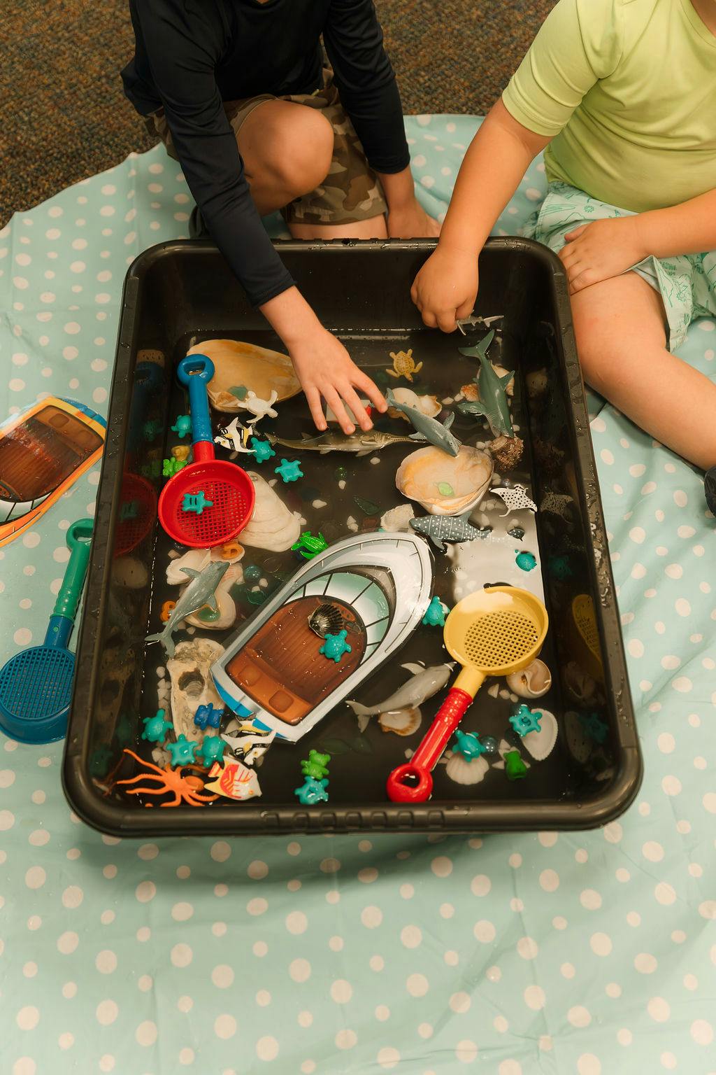People playing with toy animals and a toy boat in a water-filled plastic bin