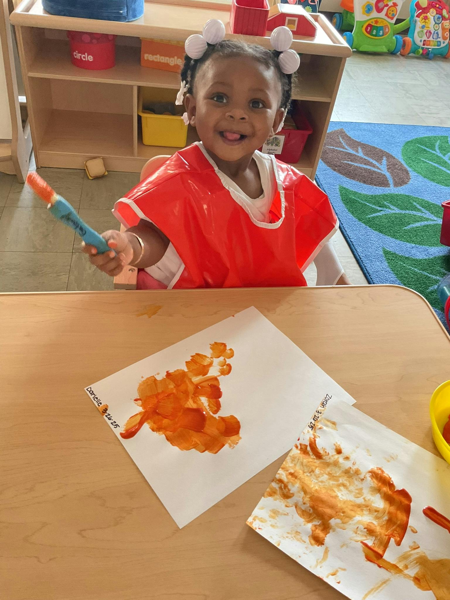 Child in a red smock holding a paintbrush over painted papers