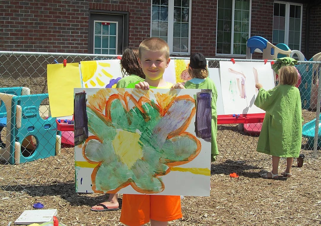 Child holding up a painting outdoors, other children painting in background