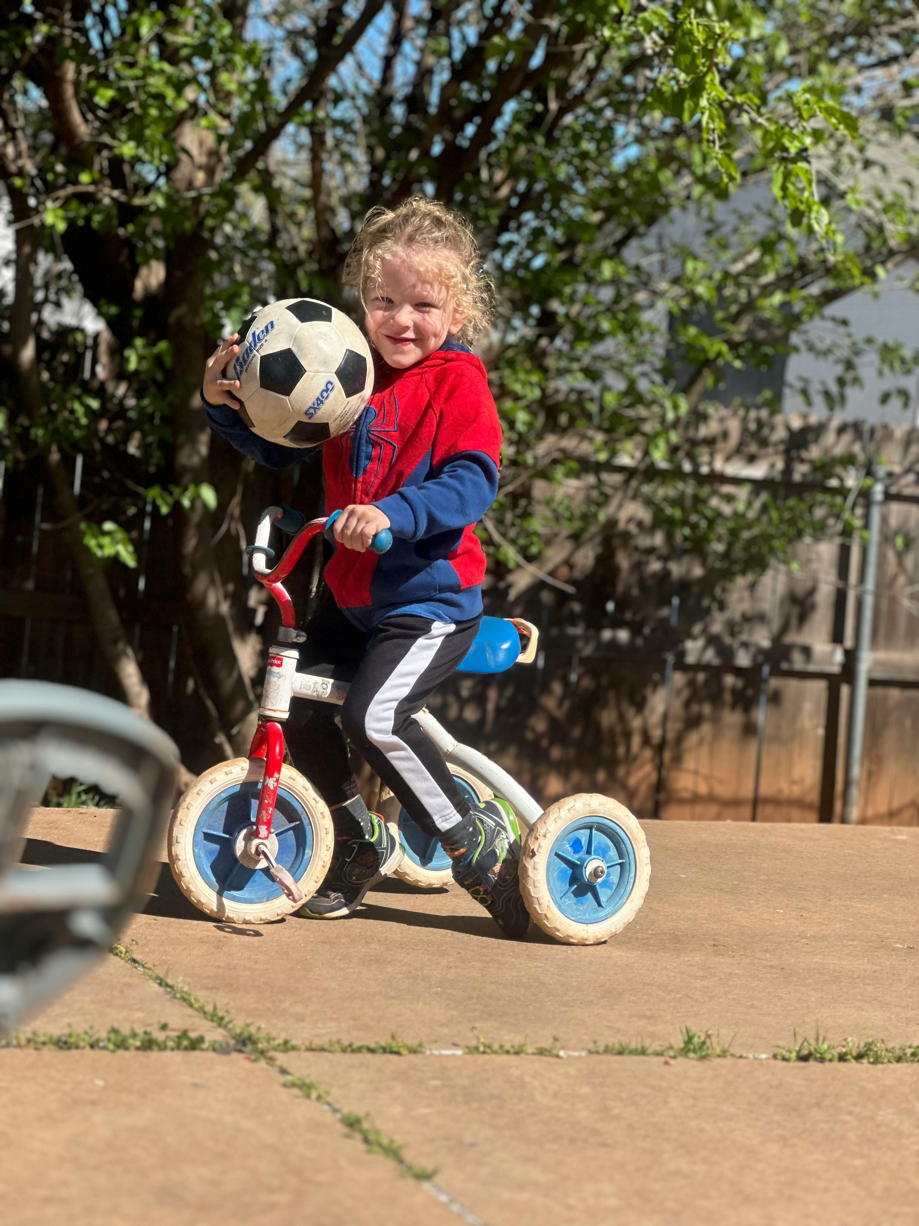 Child on a tricycle holding a soccer ball