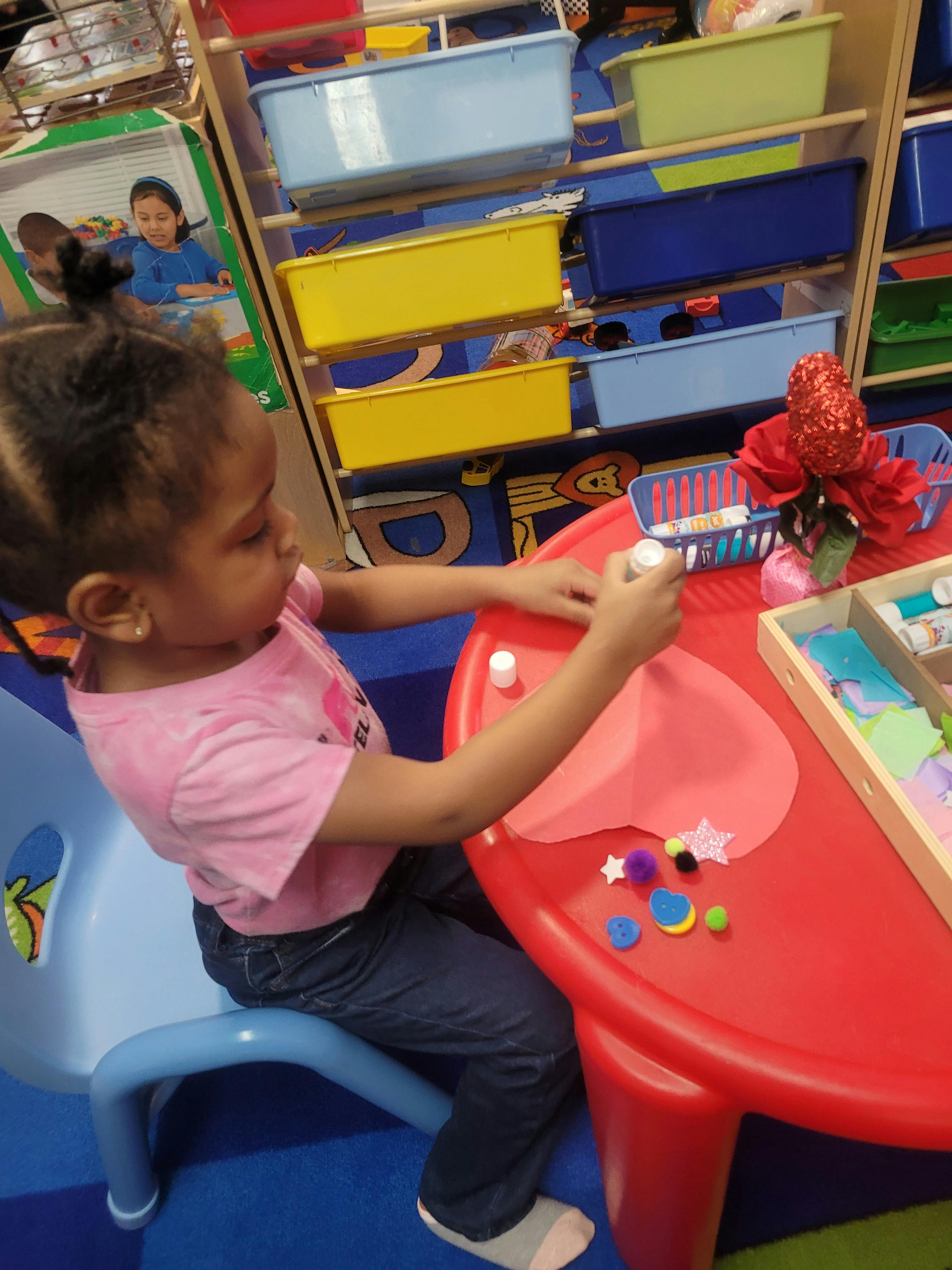 Child gluing craft items onto paper at a red table in a classroom