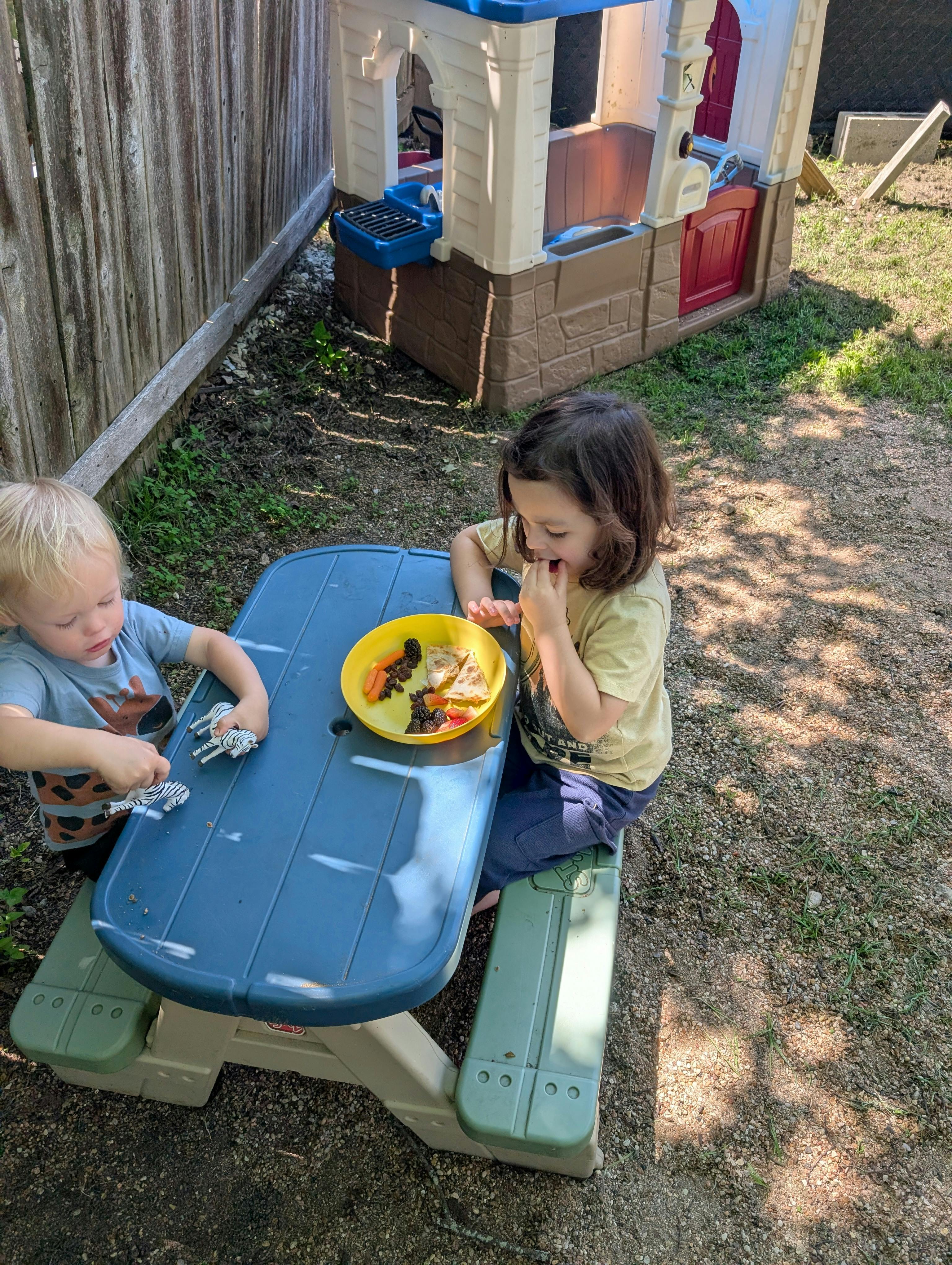 Two children at a picnic table, one eating and one playing with toys