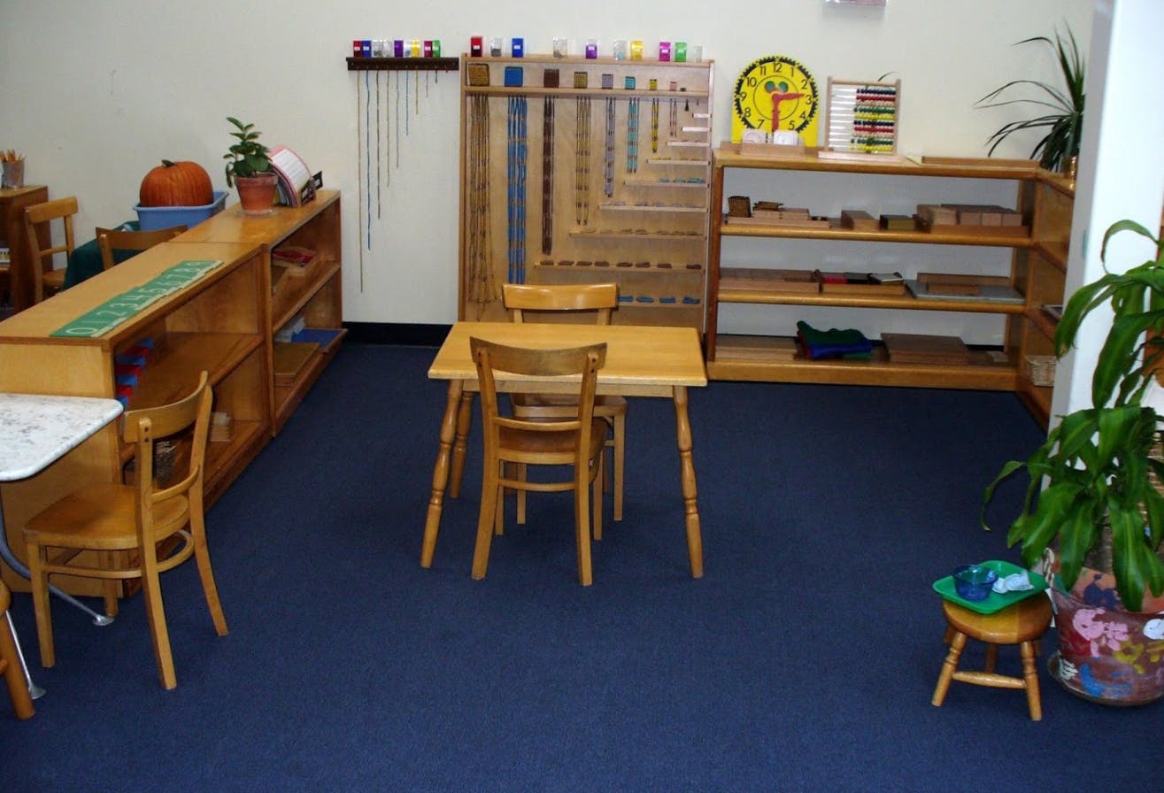 Classroom with small wooden tables and chairs, shelves with learning materials