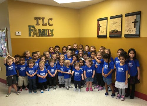 Group of children in blue shirts standing in a room with a sign that says TLC Family