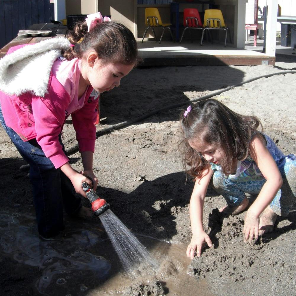 Two children playing in dirt, one spraying water from a hose
