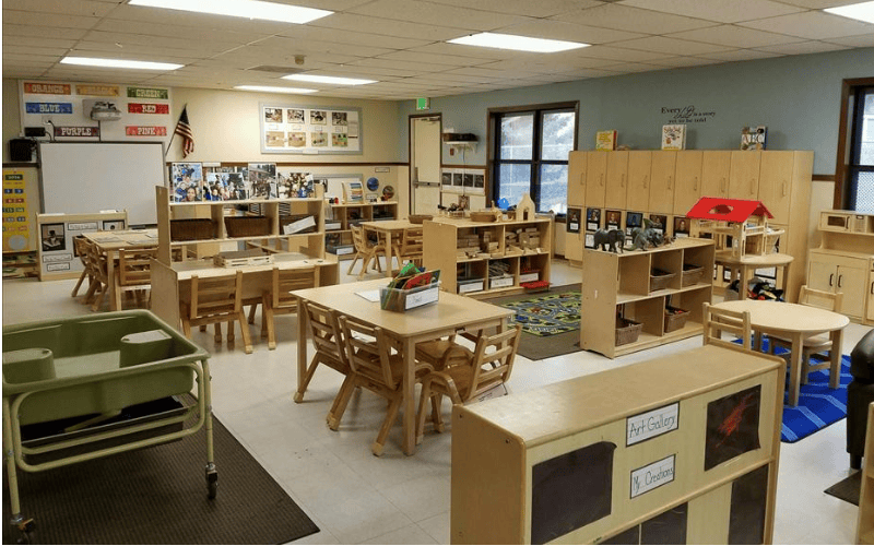 An empty classroom with child-sized tables and chairs, shelves with toys