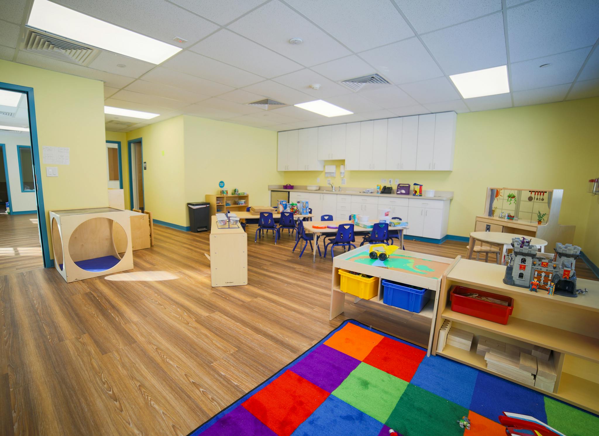 Empty classroom with small tables and chairs, play rug, and storage shelves