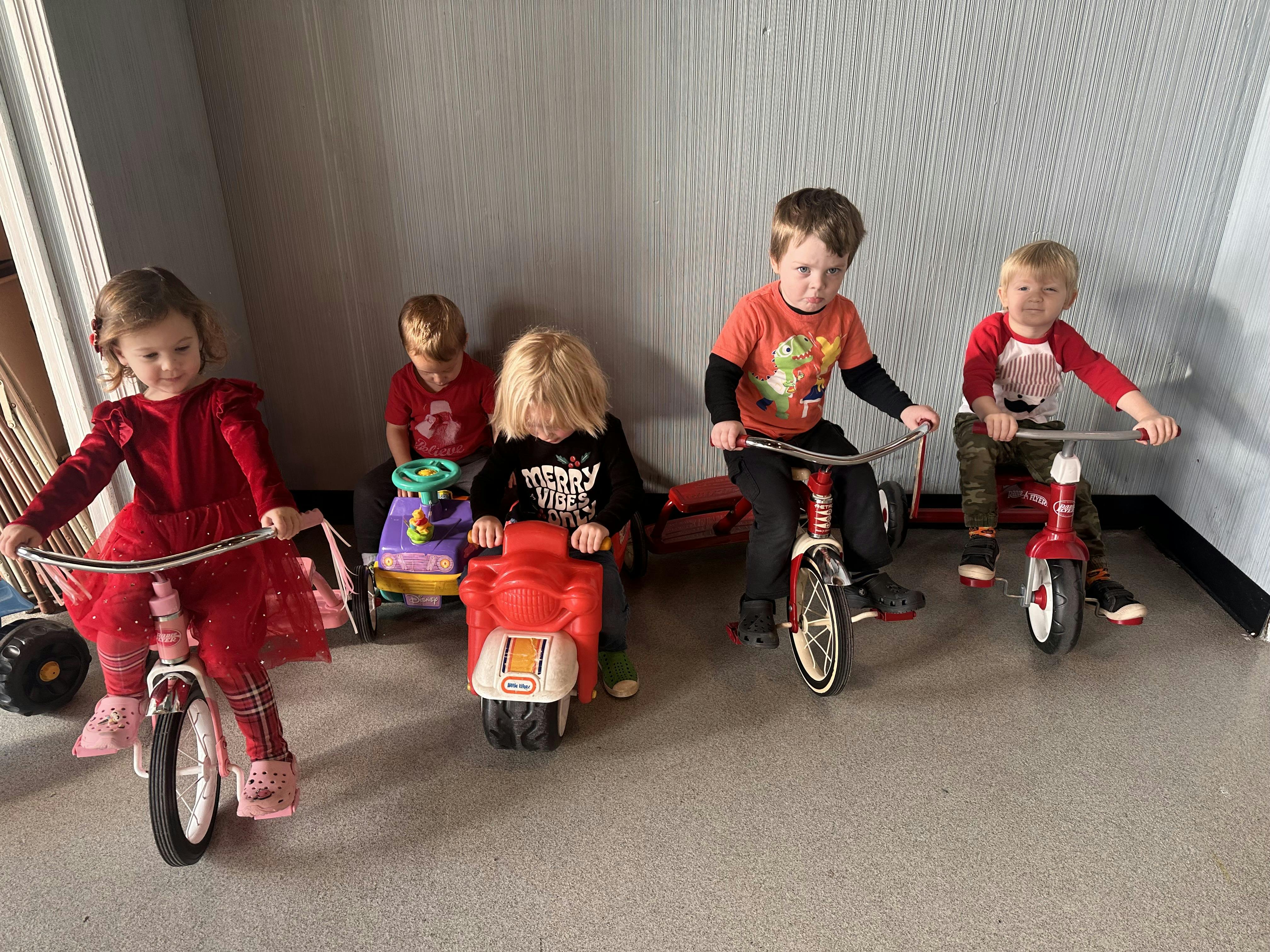 Multiple children riding tricycles and toy vehicles indoors
