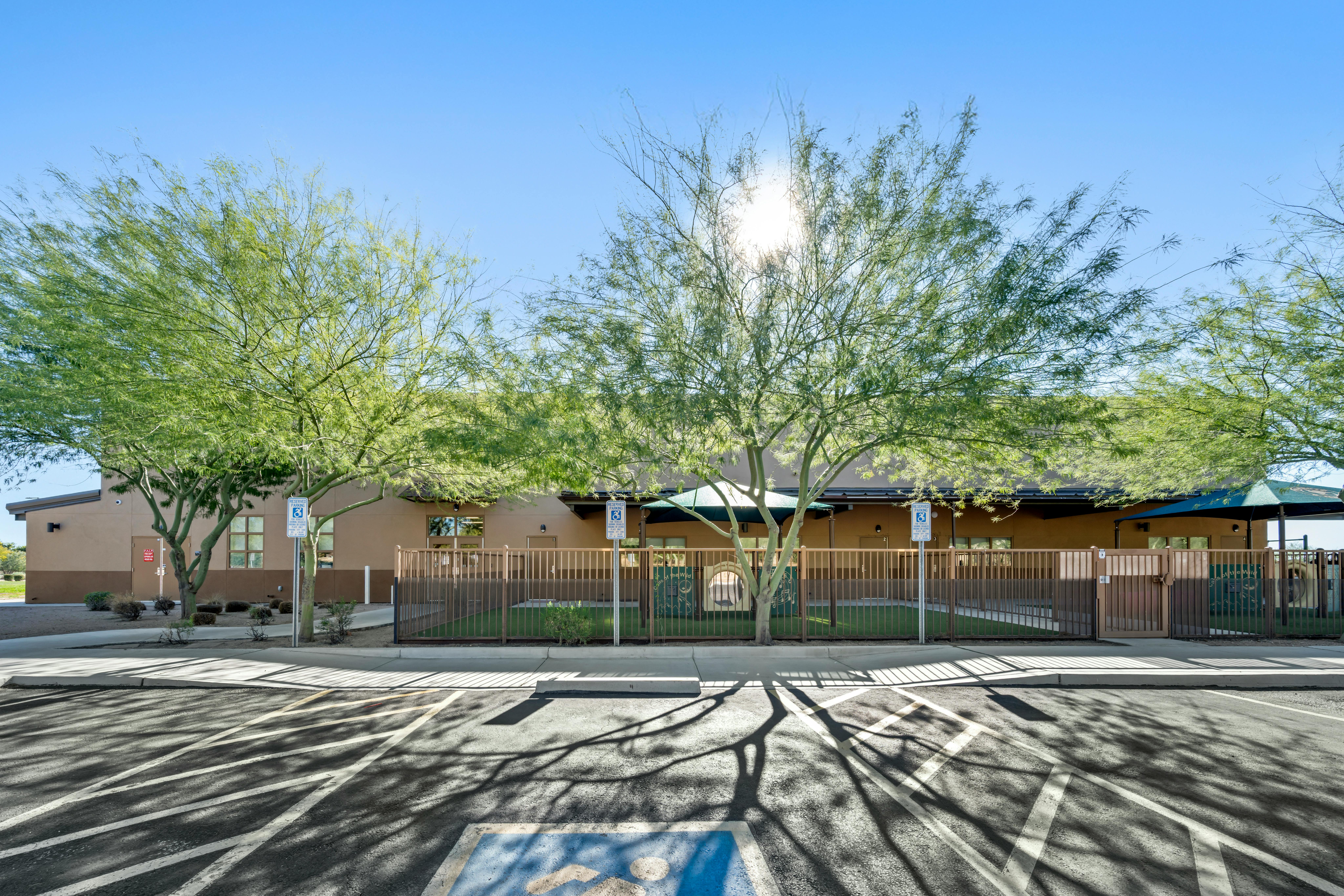 A beige building behind a brown fence and two trees, with a parking lot in front