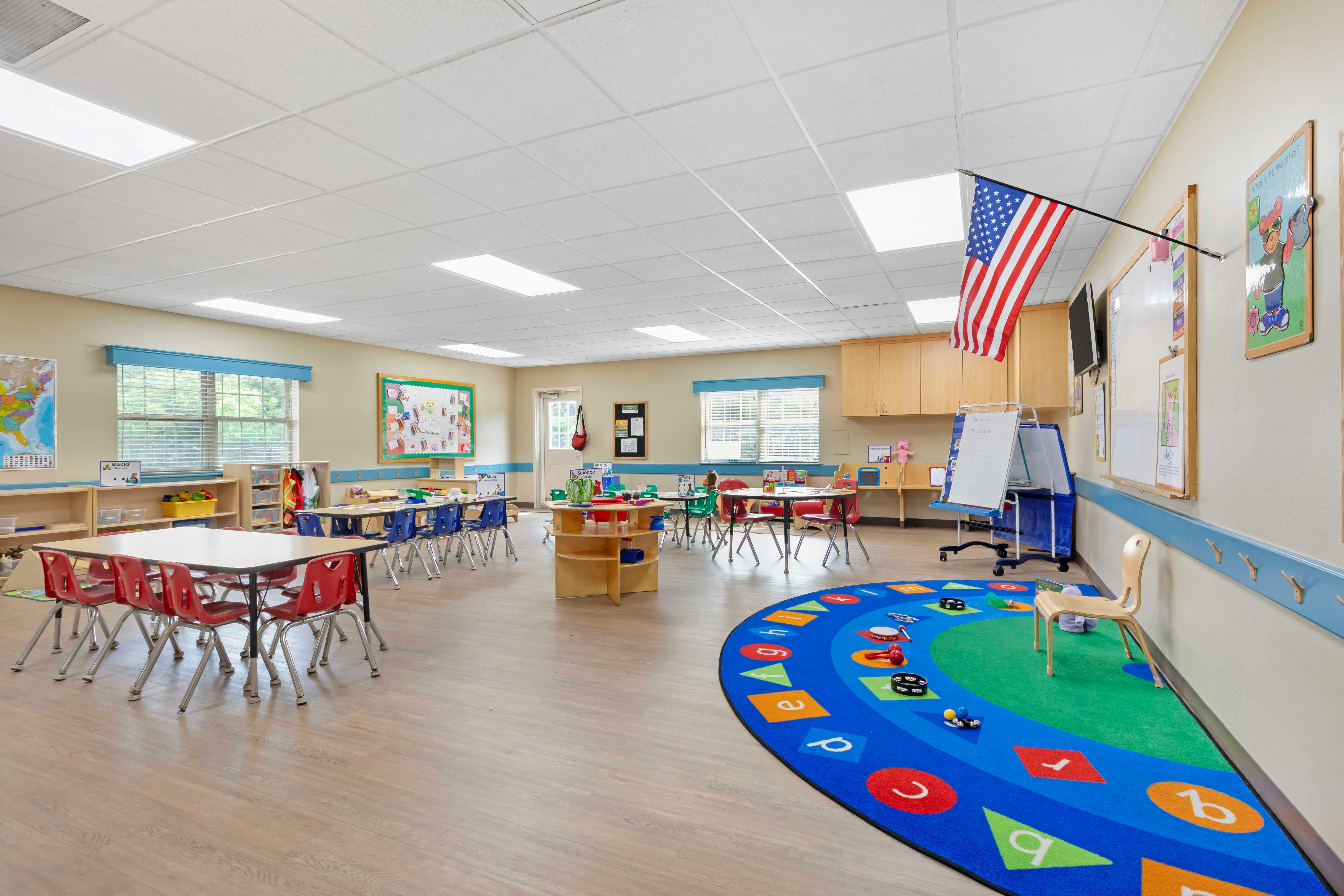 An empty kindergarten classroom with small tables and chairs, and a colorful rug