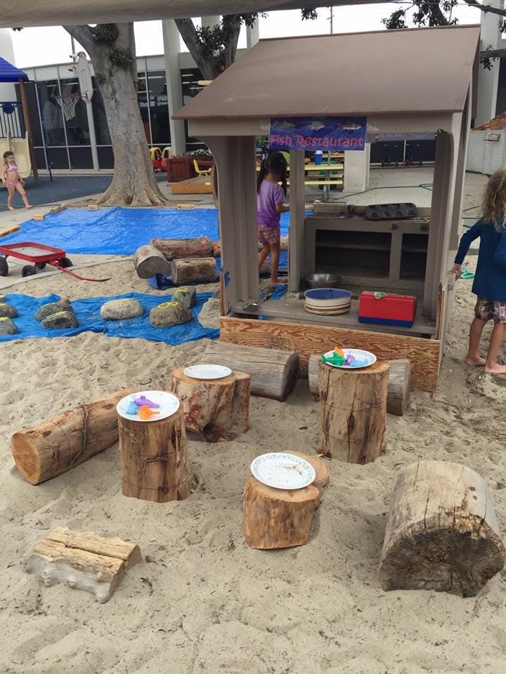 Children playing in a sand area with a small wooden playhouse and tree stumps