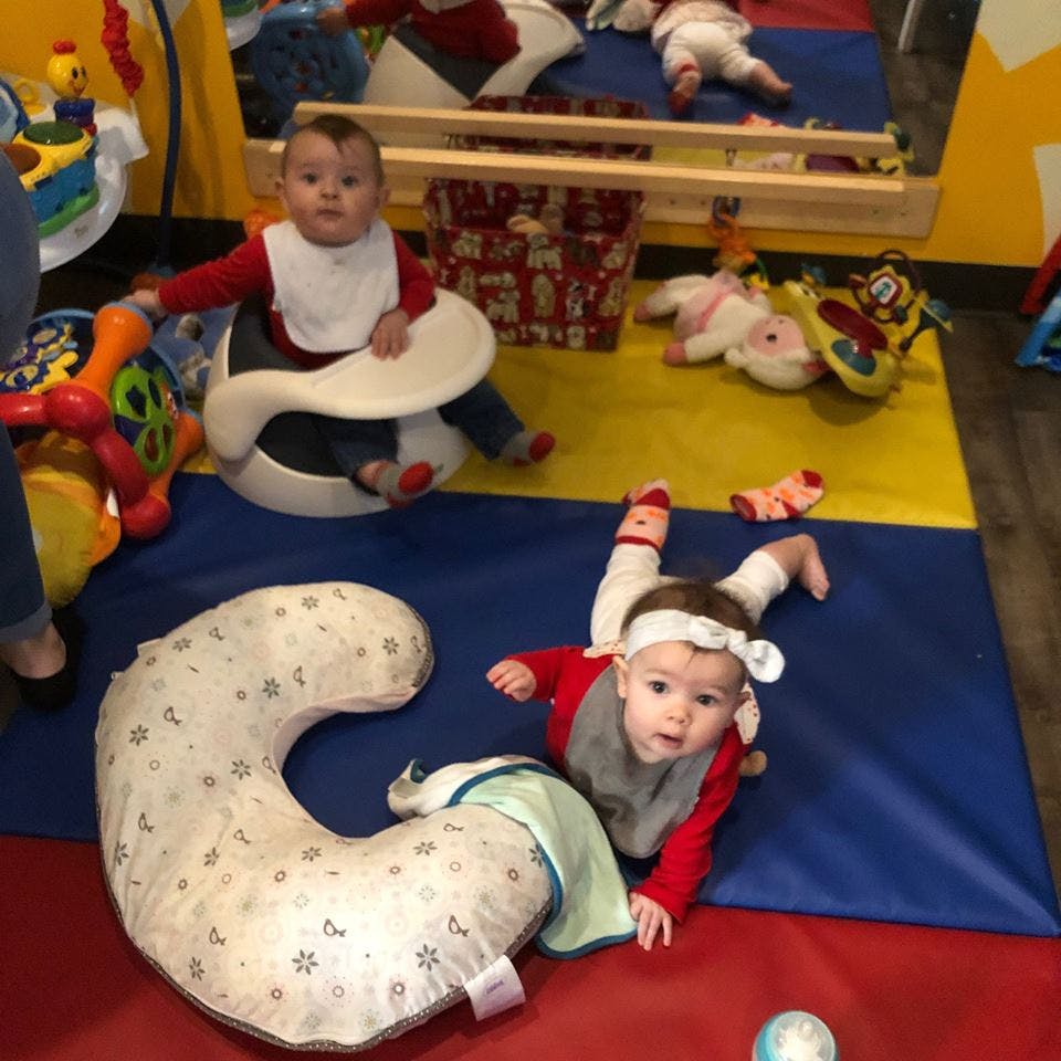 Two babies on a colorful playmat, one sitting in a seat, one on their stomach