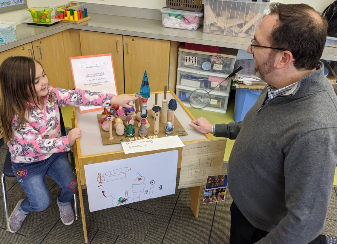Child pointing at a display of small wooden figures on a table with a person