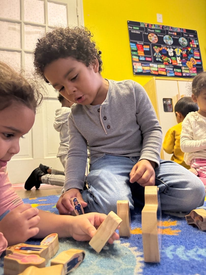 Children playing with wooden blocks and figures on a colorful rug in a classroom