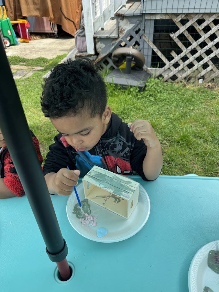 Child painting a small wooden box on a blue table outdoors