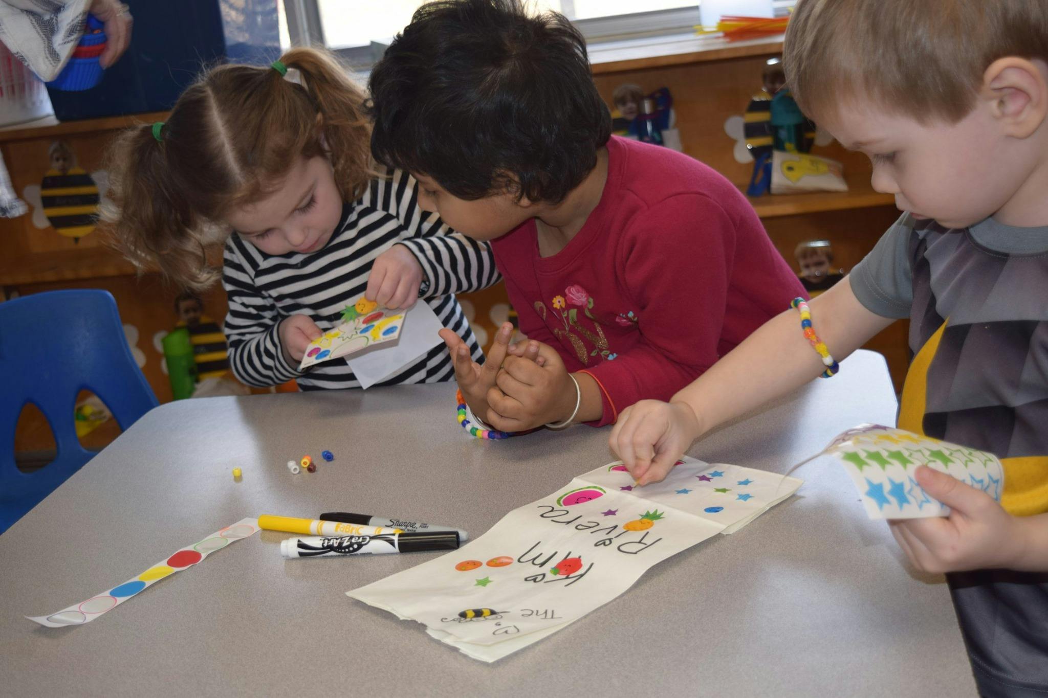 Three children at a table doing an art activity with beads and markers