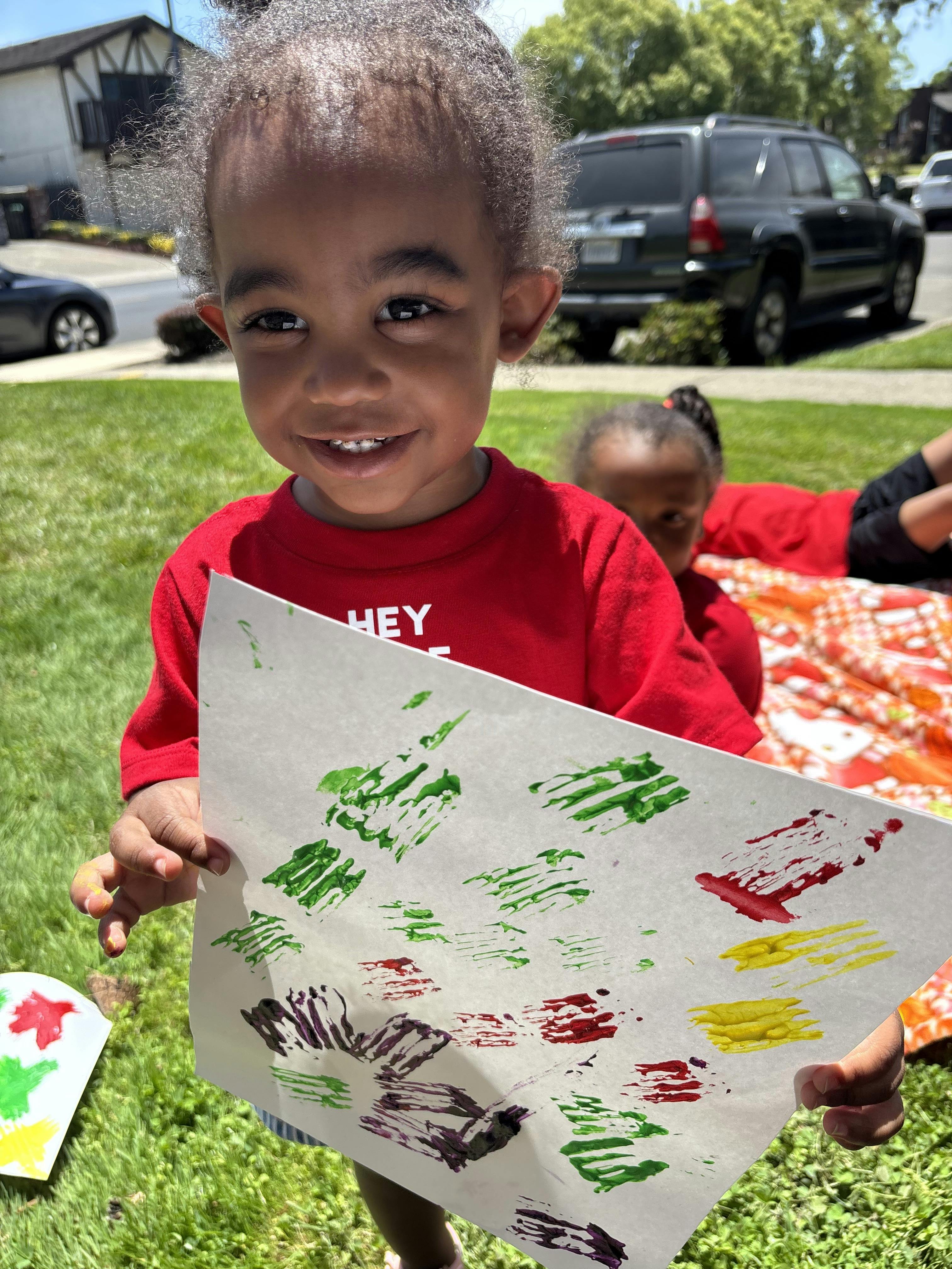 Child holding up paper with paint handprints in a yard
