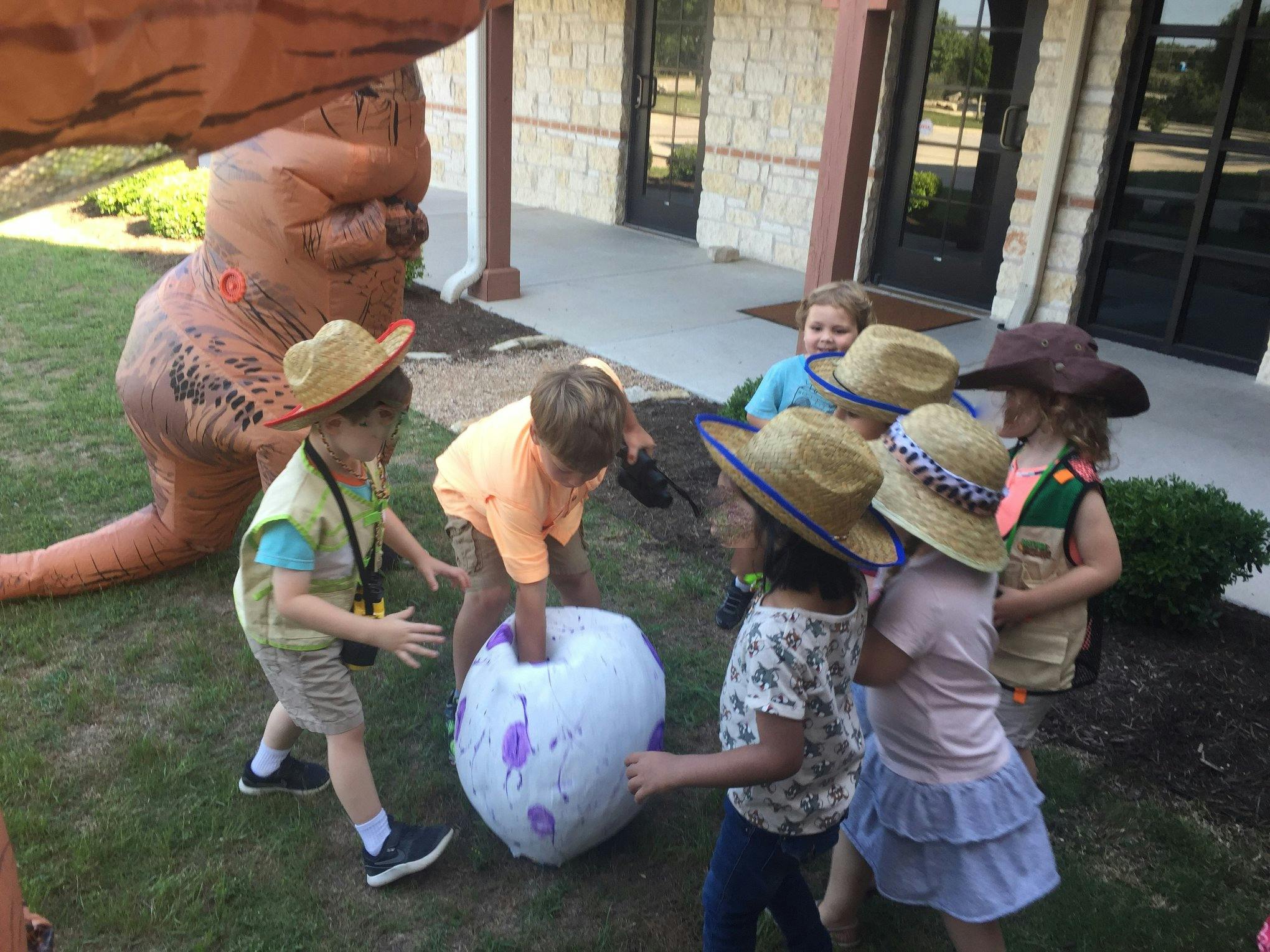 Children in hats playing with a large white object near an inflatable dinosaur