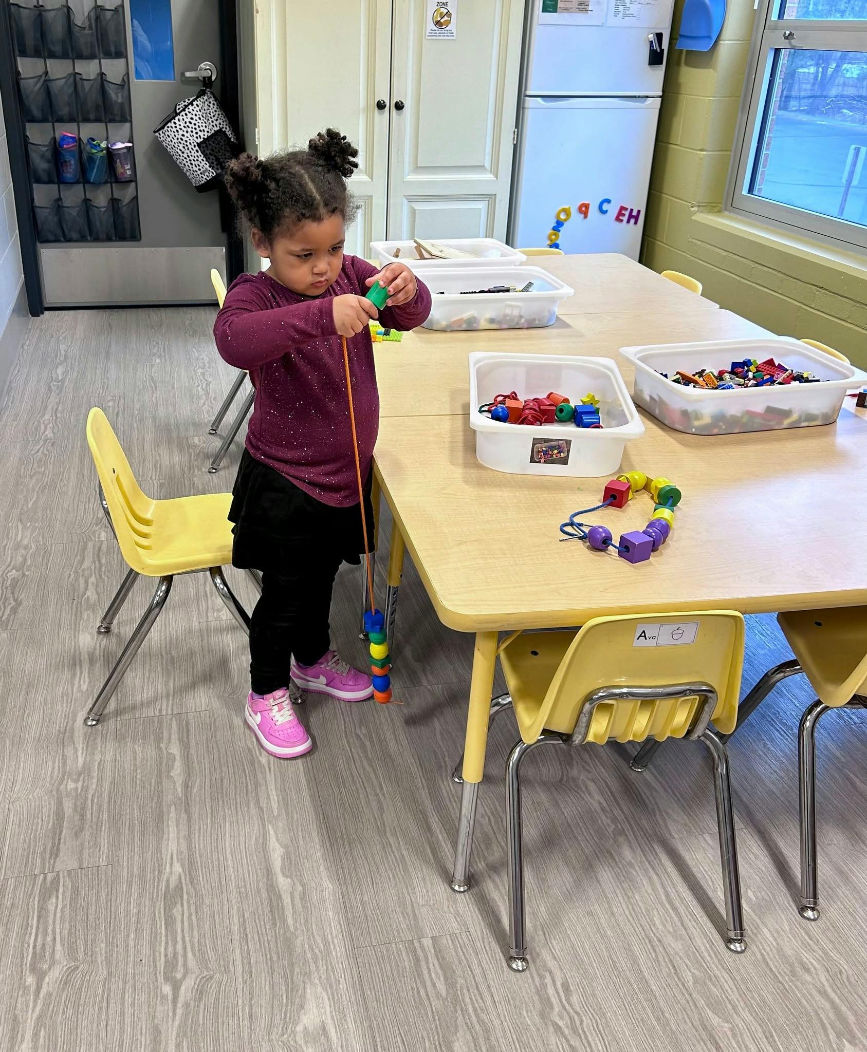 Child stringing beads at a table in a classroom
