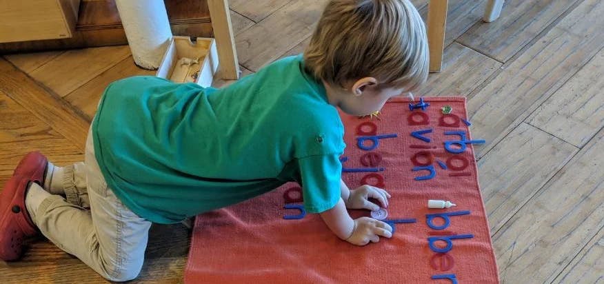 Child kneeling on floor, looking at a red mat with blue letters