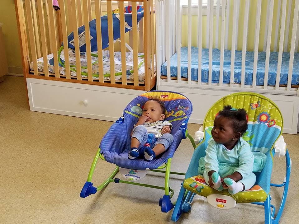 Two infants sitting in bouncer chairs in a room with cribs