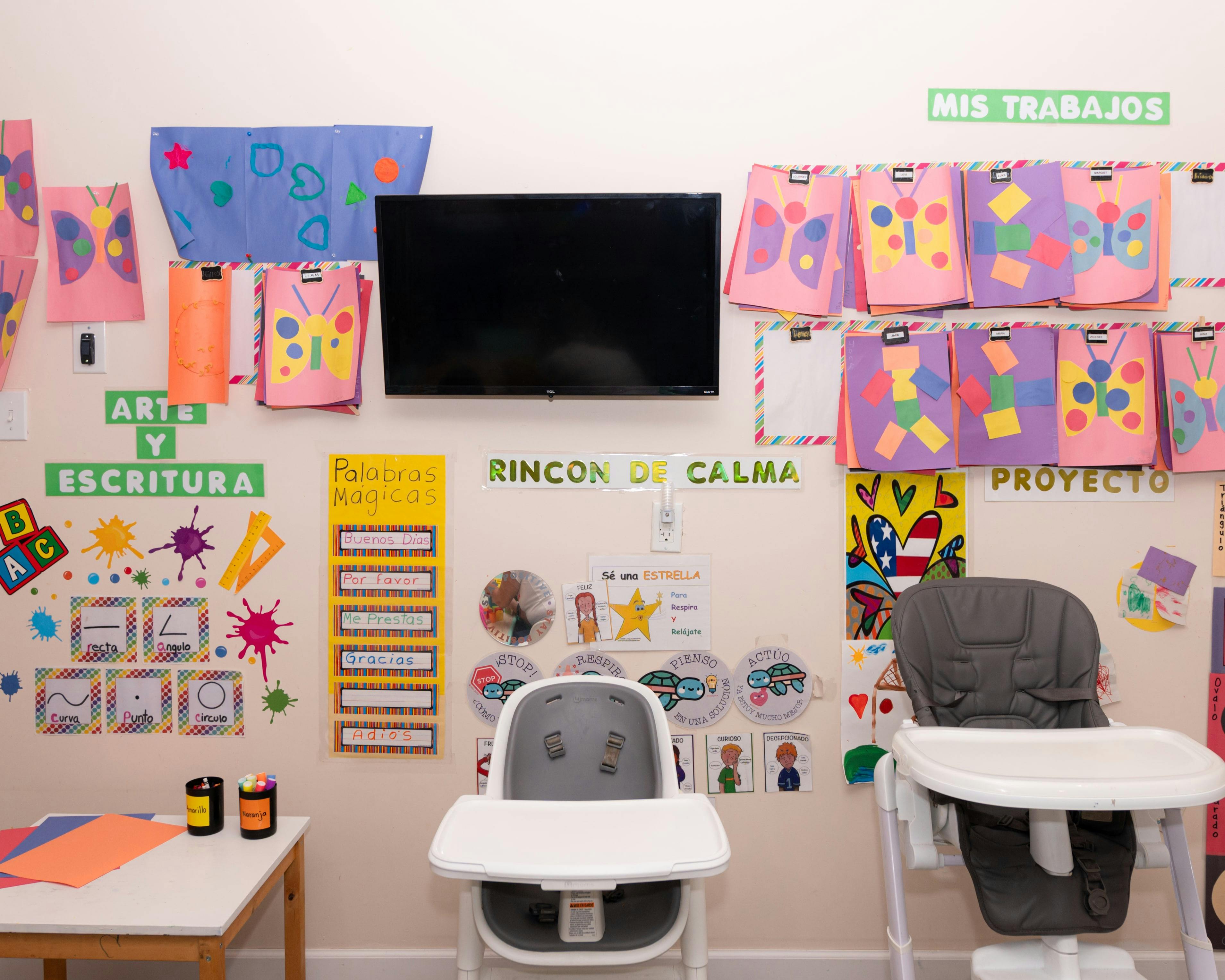 Classroom wall with children's artwork, a TV, and two high chairs