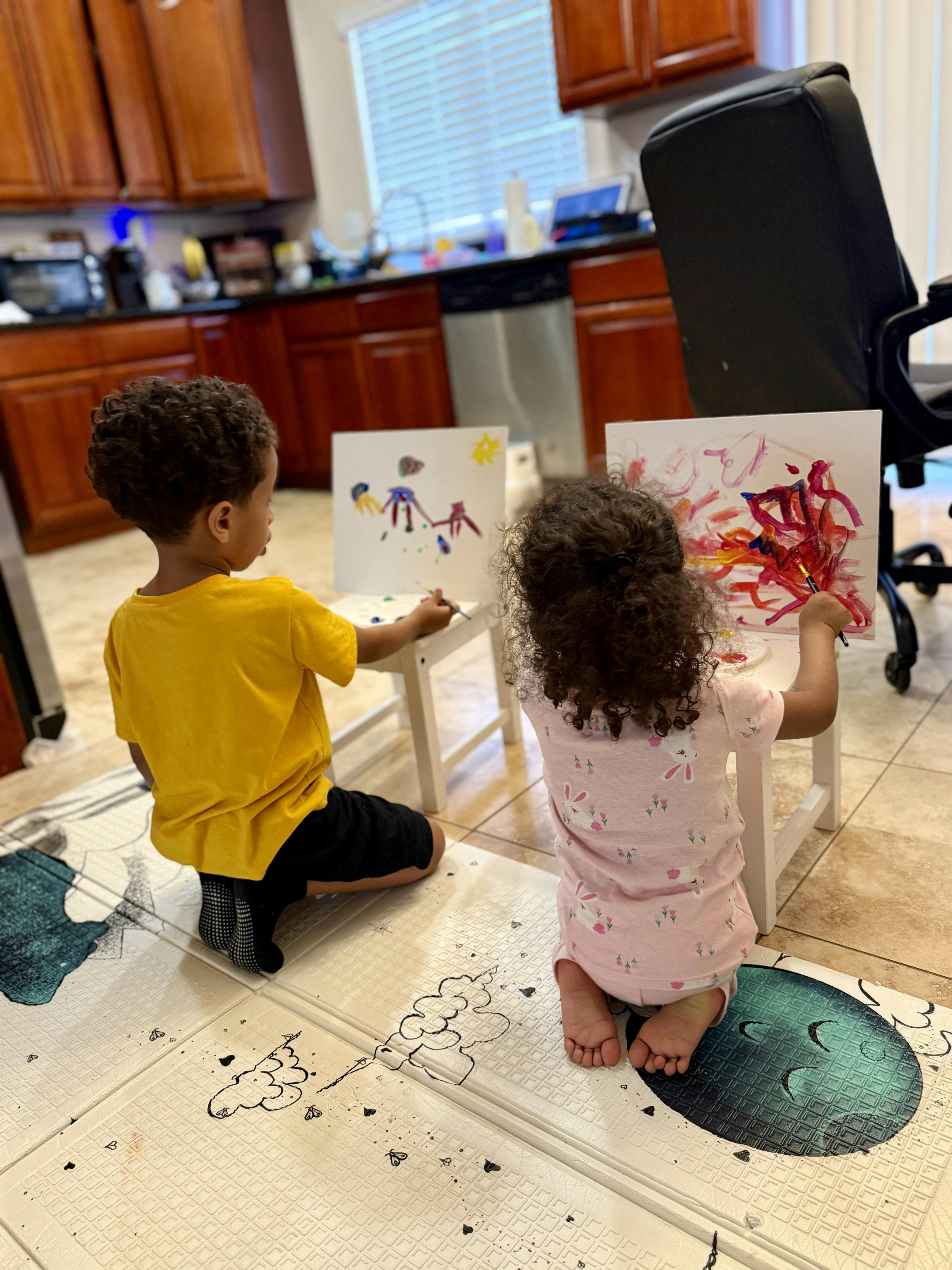 Two children kneeling on a play mat painting on small easels in a kitchen
