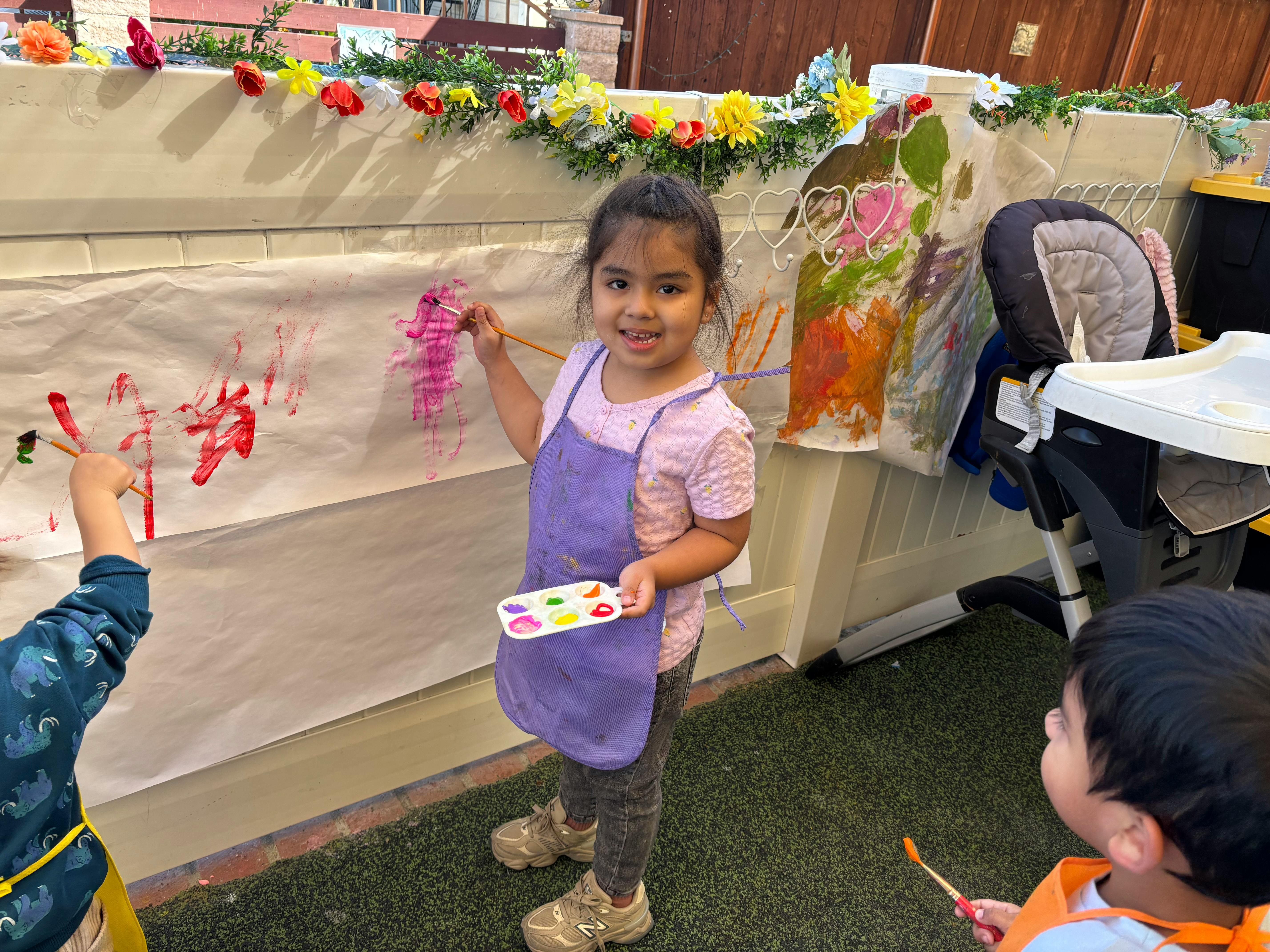 Child in apron holding paint palette, painting on paper with other children
