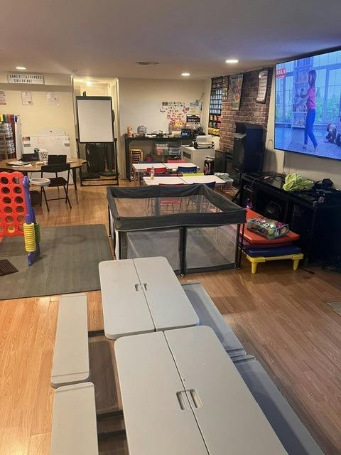 Childcare playroom with small tables, chairs, playpen, and wall-mounted screen