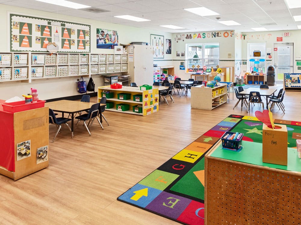 Empty classroom with small tables and chairs, shelves, and a colorful rug