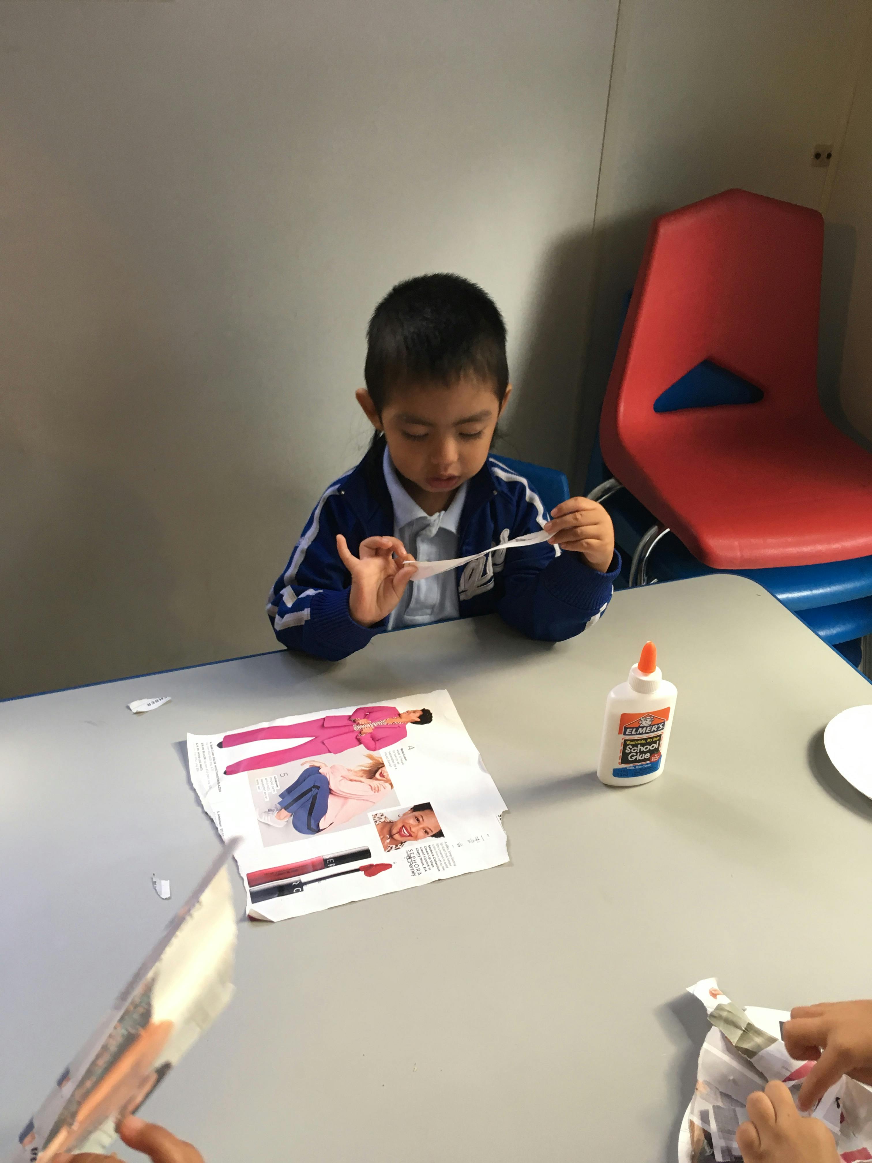 Child sitting at a table, holding a piece of paper, with a magazine and glue on the table