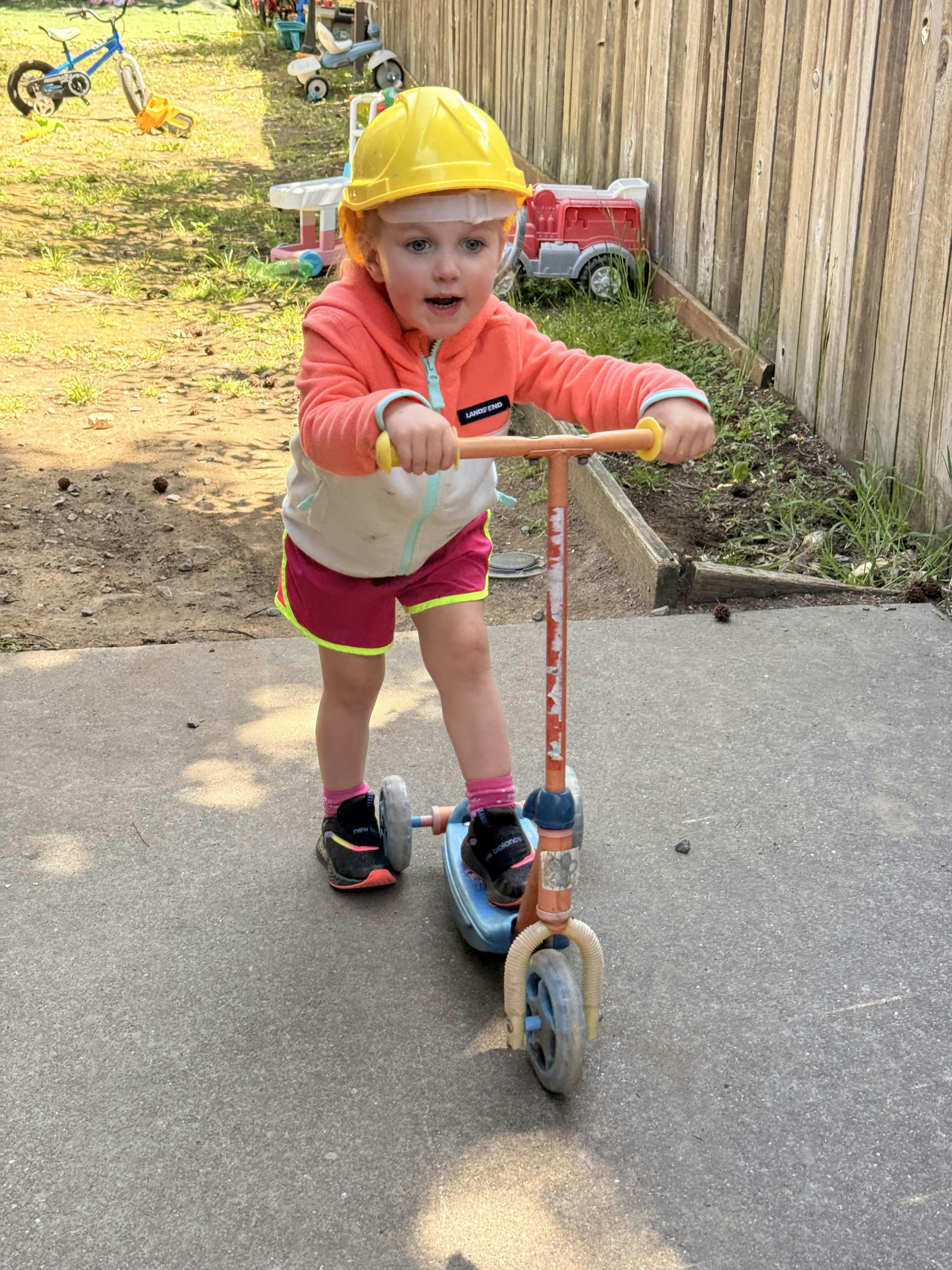 Child wearing a yellow hard hat riding a scooter