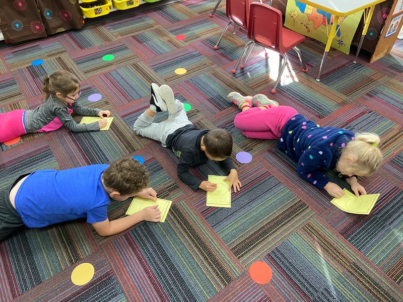 Four children lying on a carpeted floor writing on yellow paper