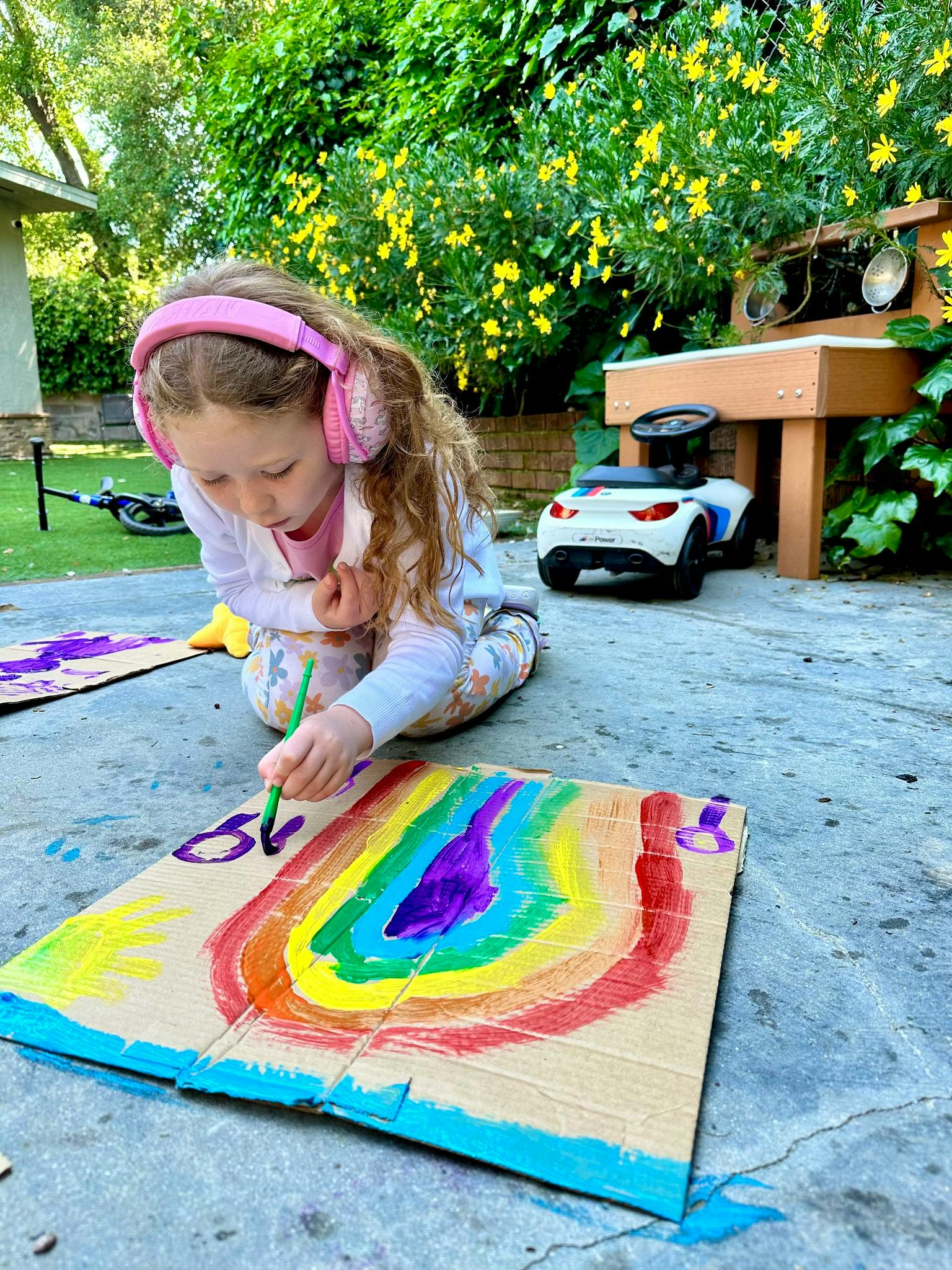 Child wearing pink headphones painting a rainbow on cardboard outdoors