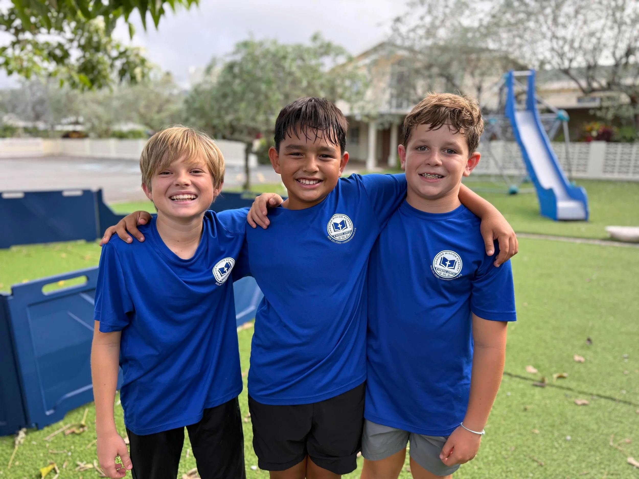 Three children in blue school shirts with arms around each other at a playground