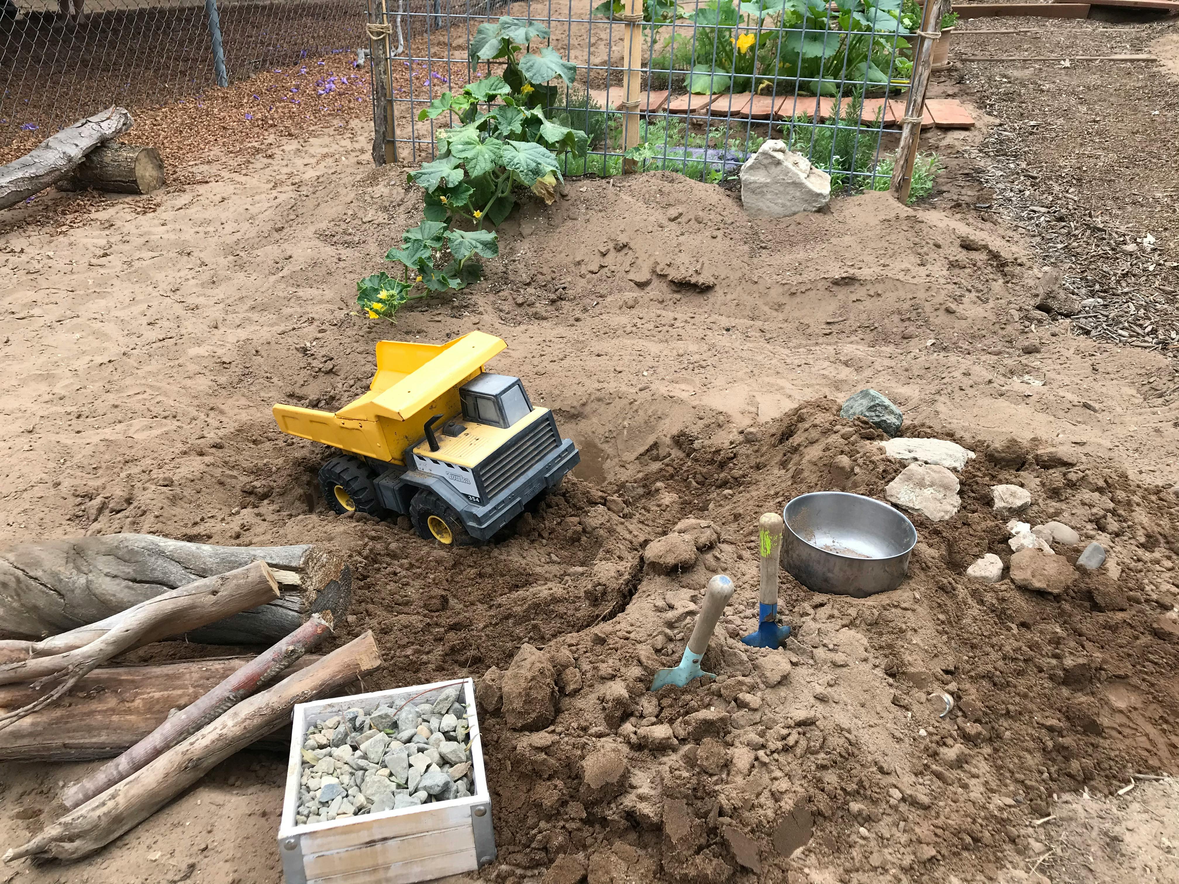 Toy dump truck in a pile of dirt with a metal bowl and small shovel