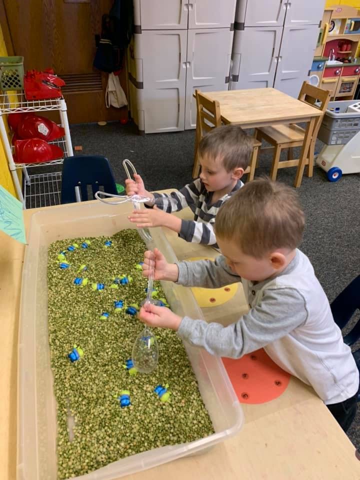 Two children playing with a sensory bin filled with green material and small toys
