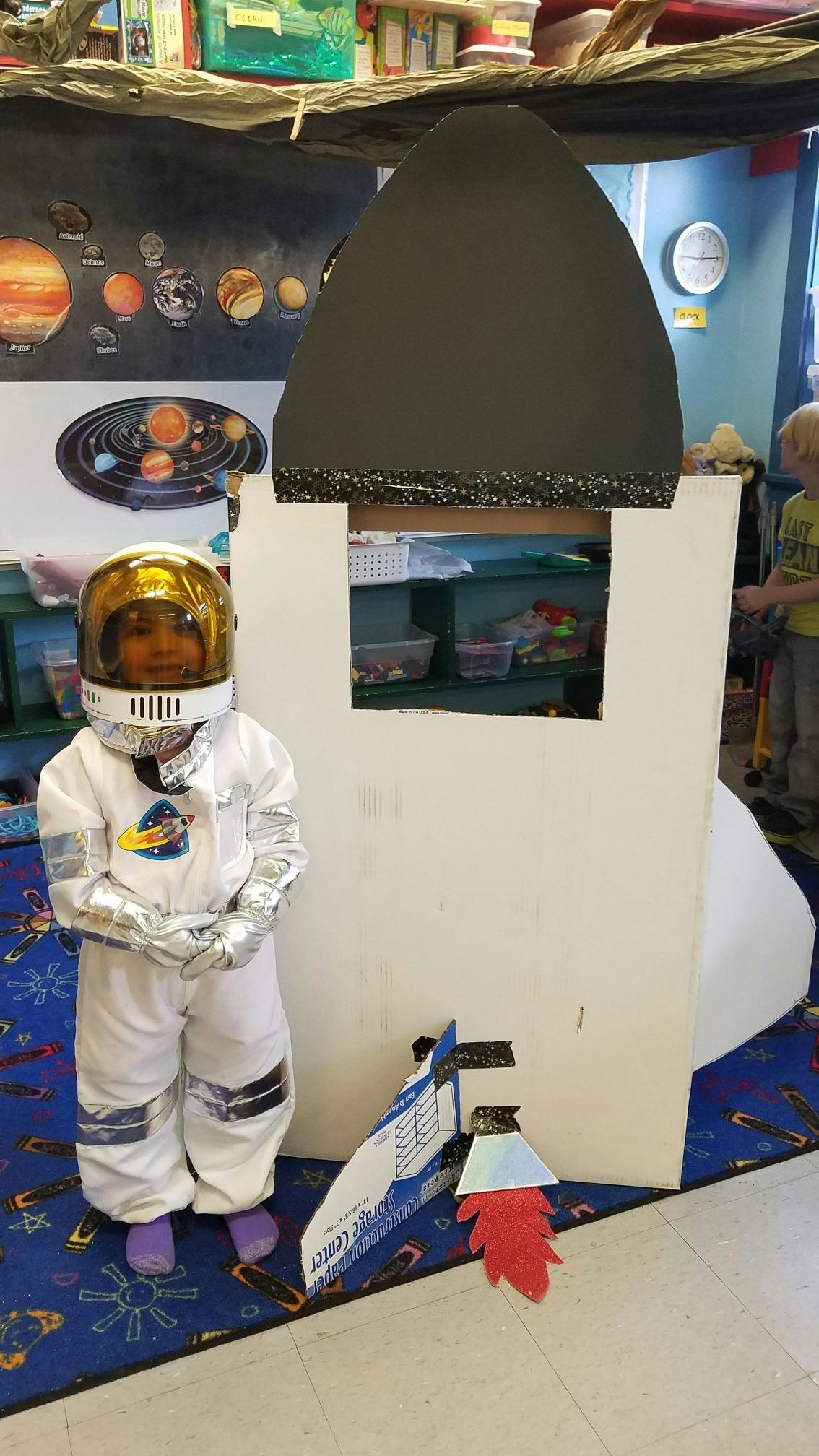 Child in astronaut costume standing next to a cardboard rocket