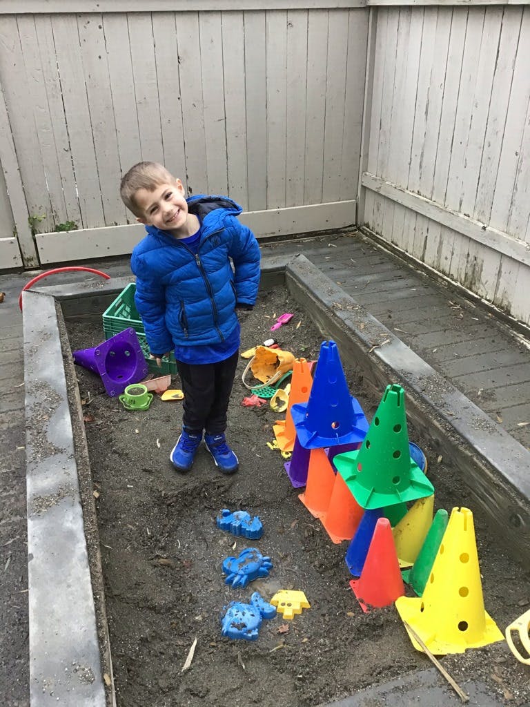 Child in a blue jacket standing in a sandbox with colorful toys